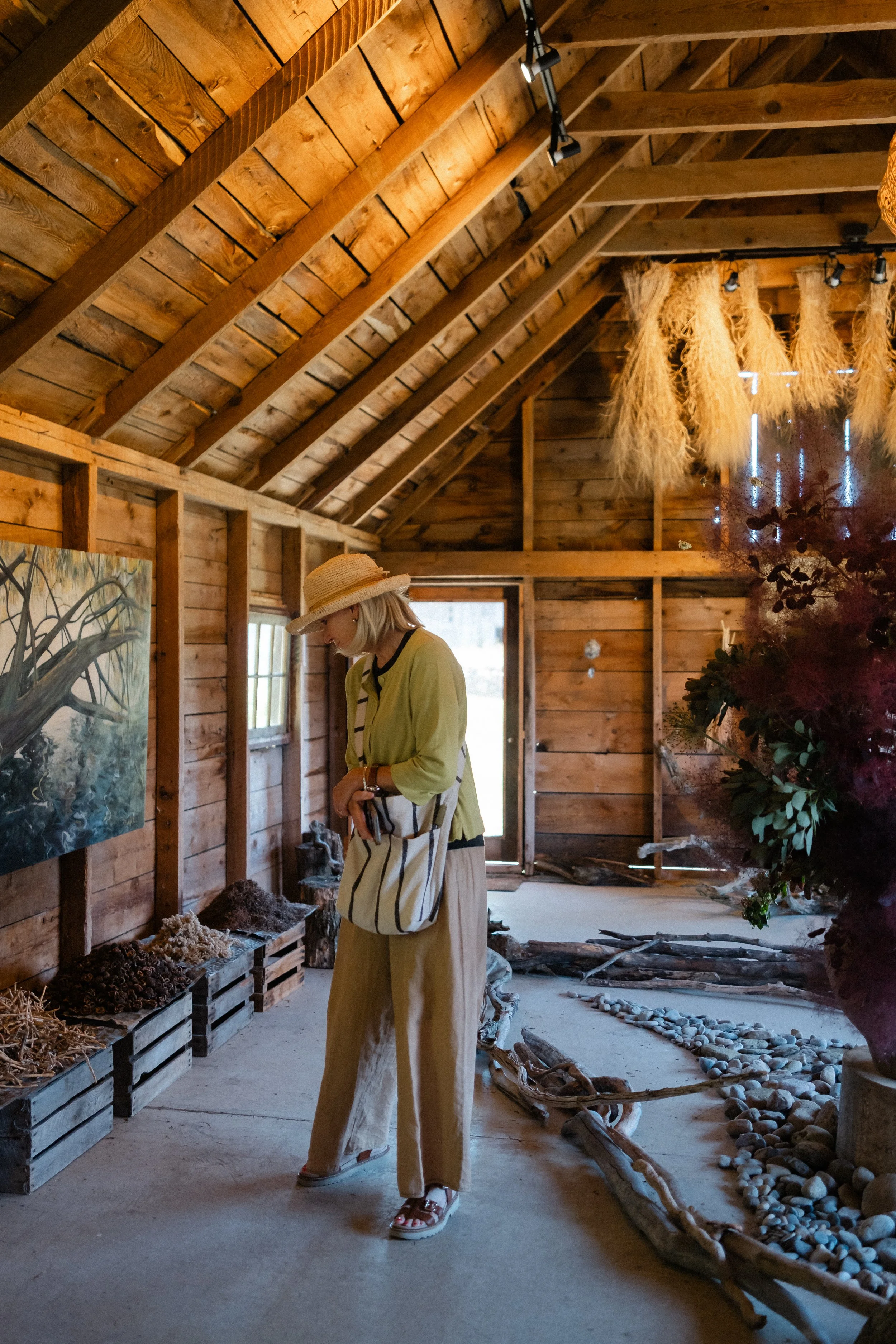 A woman wearing a straw hat and yellow jacket looking at display of natural materials and found objects as part of an installation for Flowering Wild inside a wooden building with high ceilings.