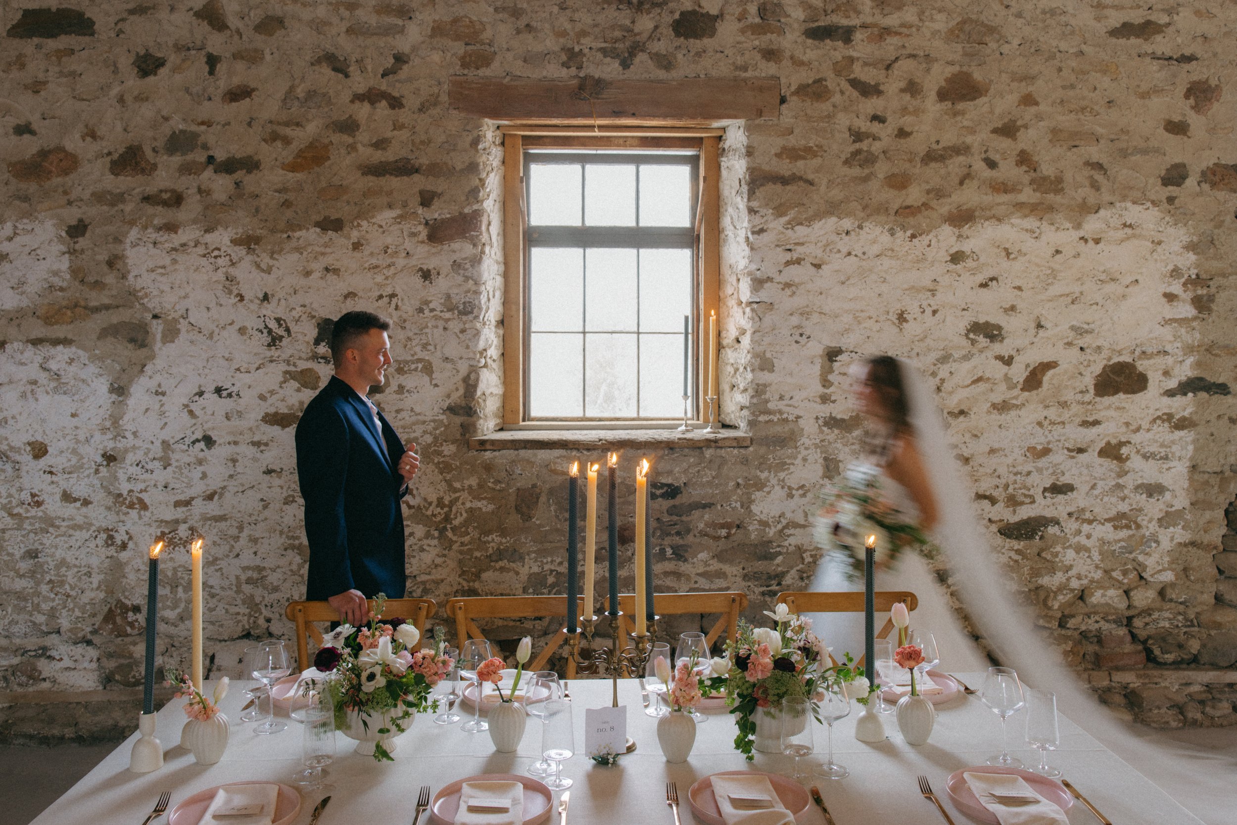 An ethereal, soft-focus documentary photograph of a bride and groom behind an elegant tablescape, featuring glowing beeswax candles and artisanal floral design.