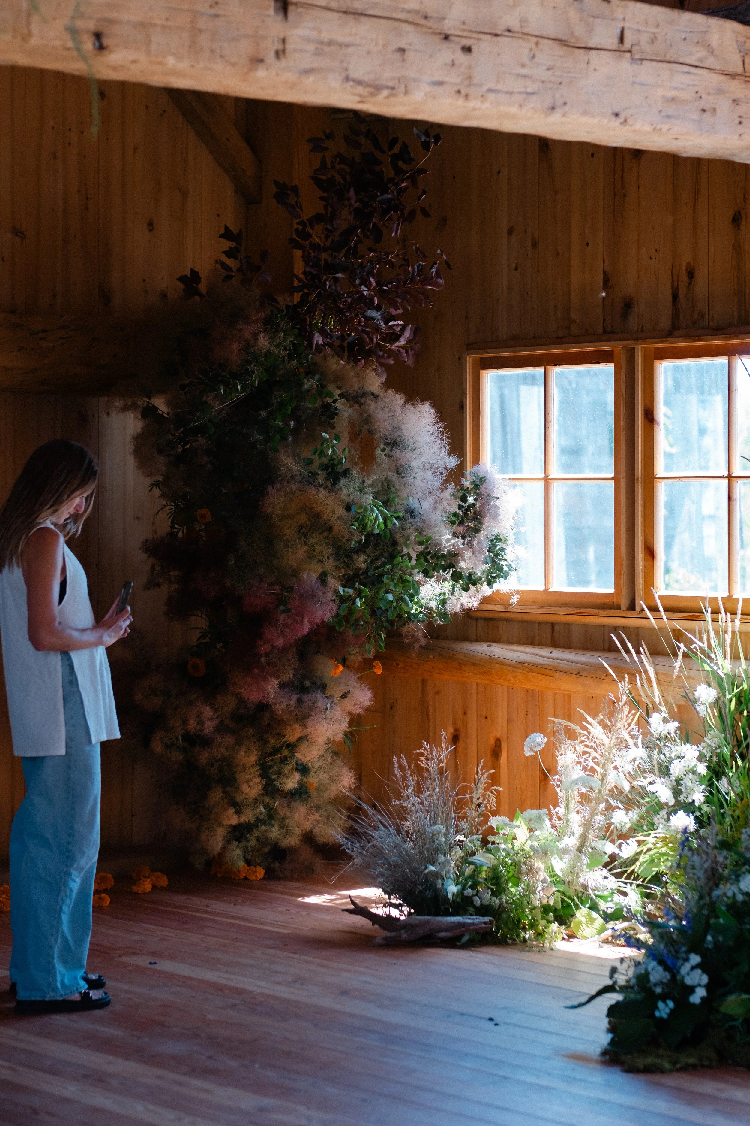 A woman wearing a white top and blue jeans standing inside a wooden barn taking photos of a floral installation as part of Flowering Wild and standing in front of a large window providing natural light.