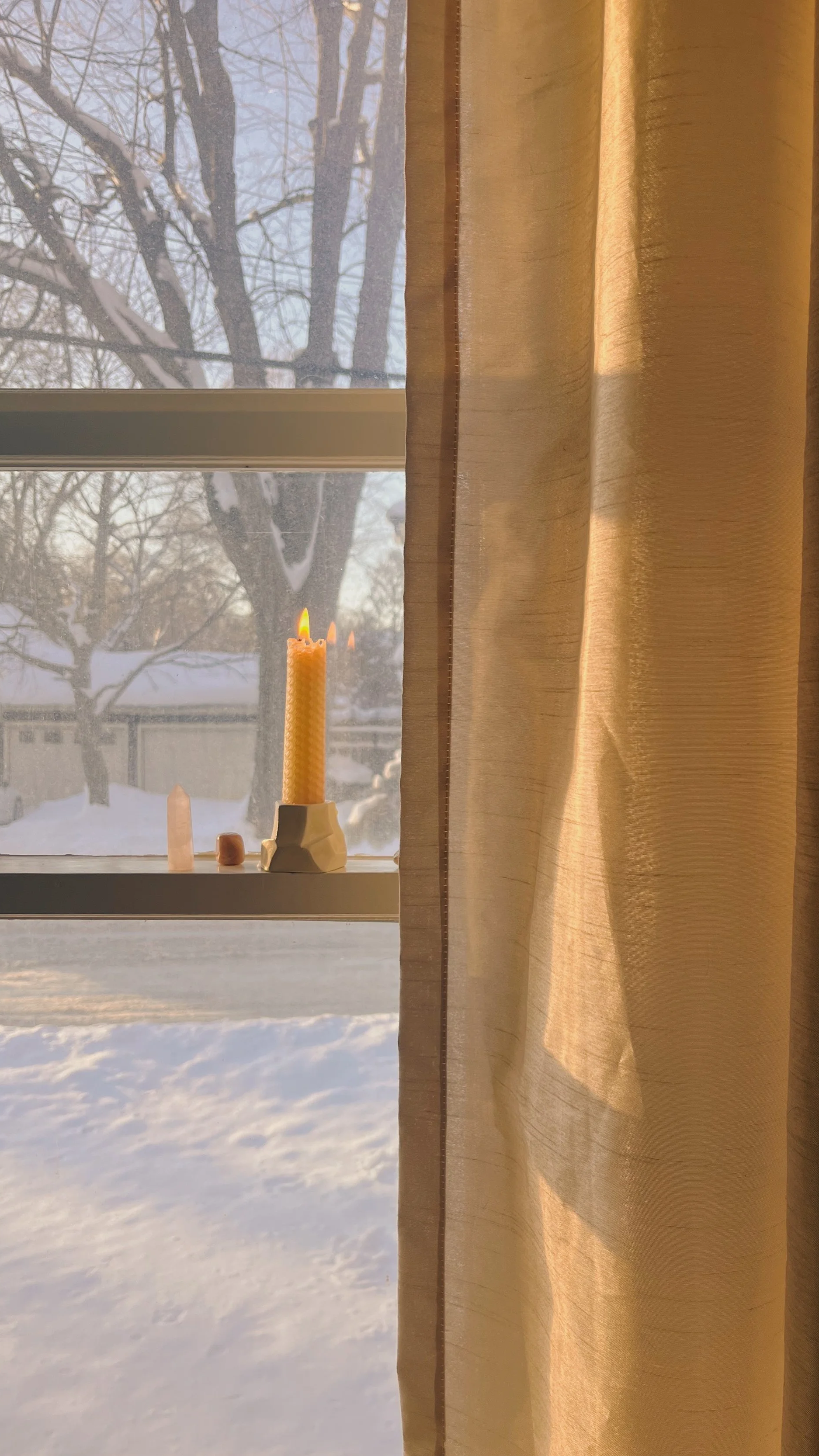 A lit natural hand-rolled beeswax taper candle burning on a window sill next to crystals, framing a bright, snowy winter scene through the glass beside soft gold curtains.