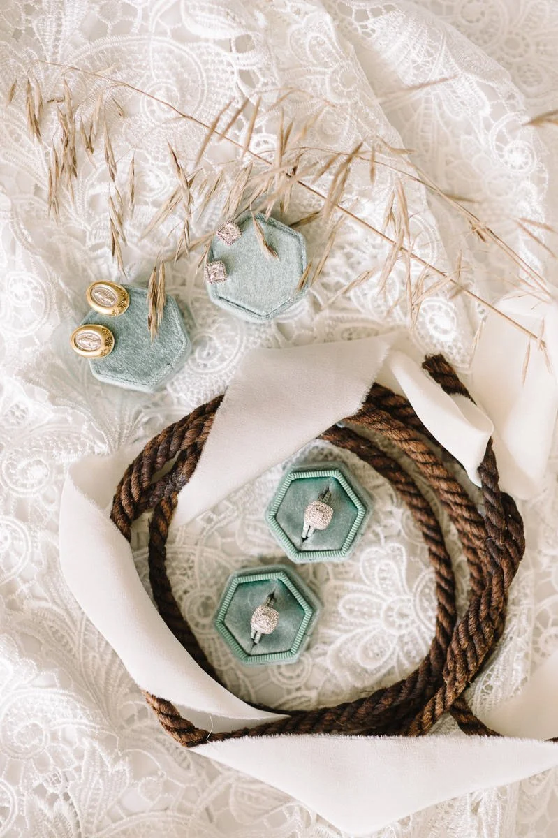 Jewelry gift set featuring diamond rings in hexagonal boxes, pair of earrings in a fuzzy box, all arranged on lace fabric with decorative dried grasses and a brown rope, presented in a white gift bag.