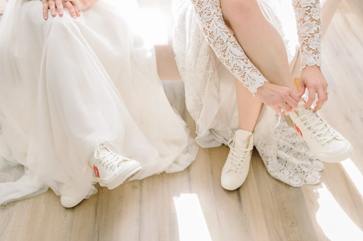 Two women in wedding dresses sitting and tying their white sneakers.