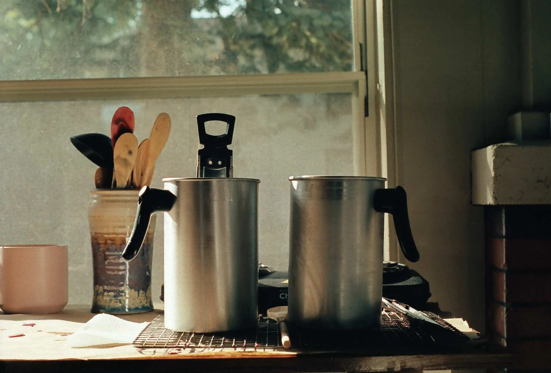Studio counter with two silver pots, utensils in a jar, and a pink mug.