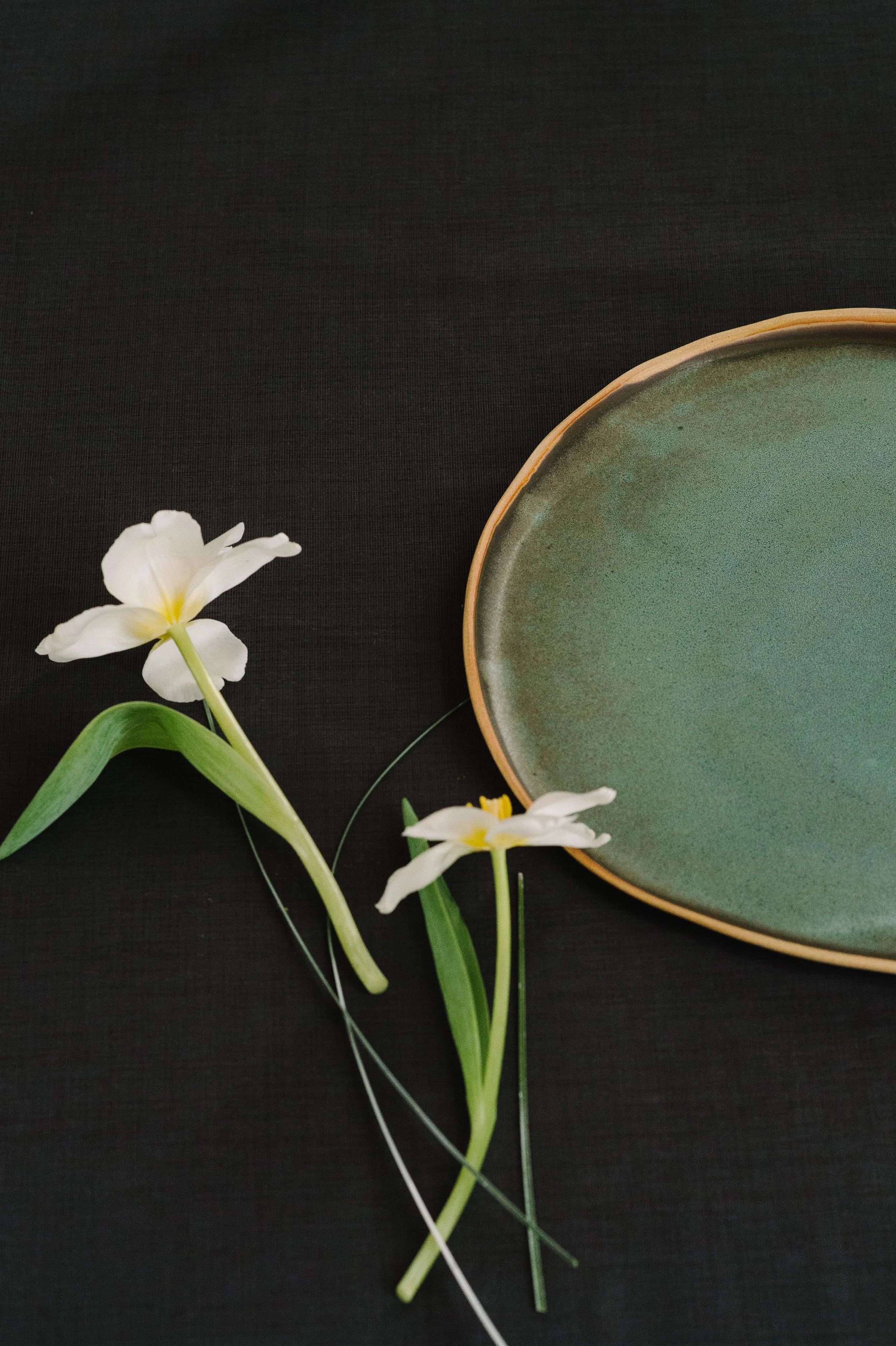 A minimalist flat lay of a handmade green ceramic plate paired with two delicate white flowers and green stems on a textured black linen surface, representing intentional design and artful tablescapes.