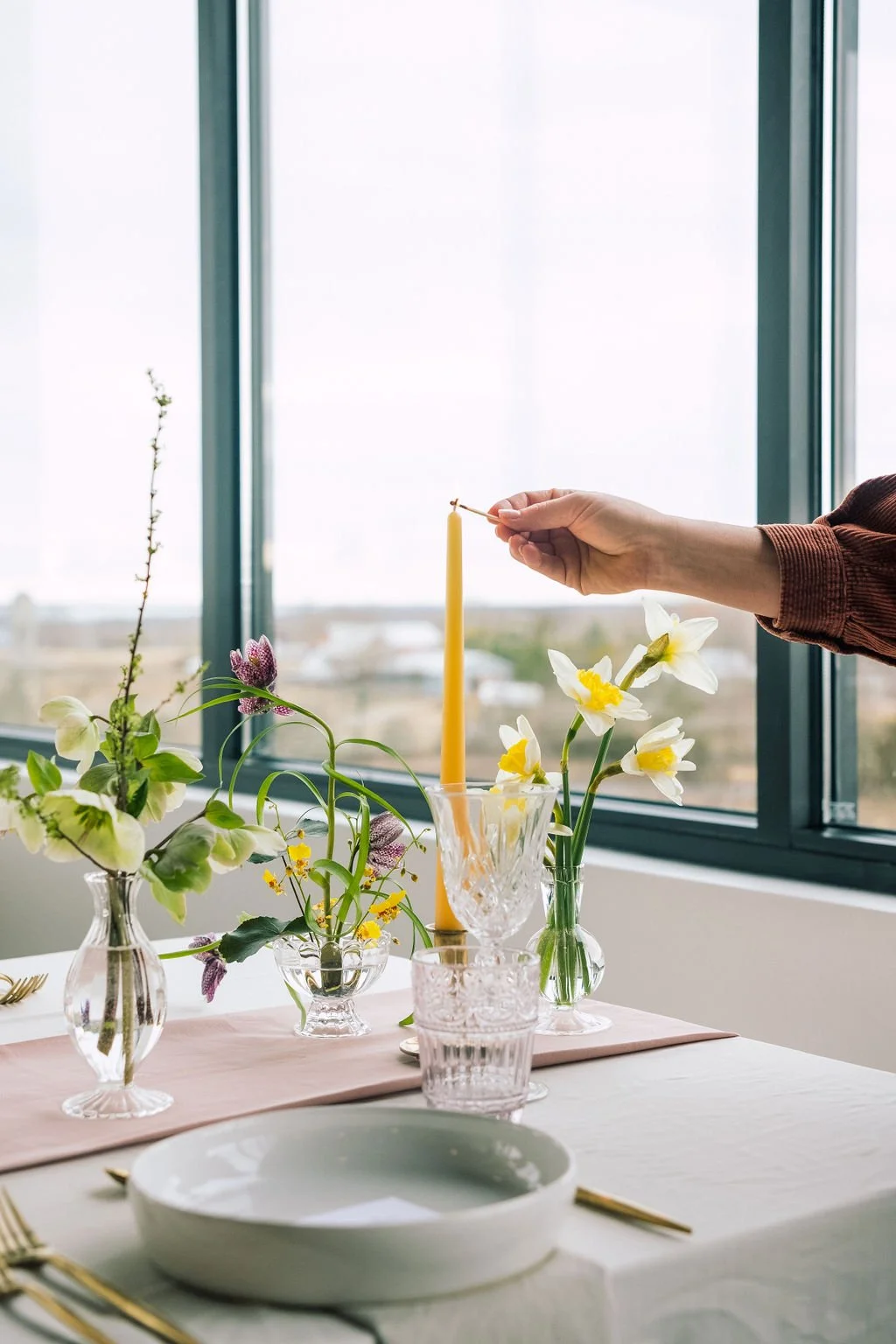 A person lighting a yellow candle on a decorated table with flower arrangements near a large window.
