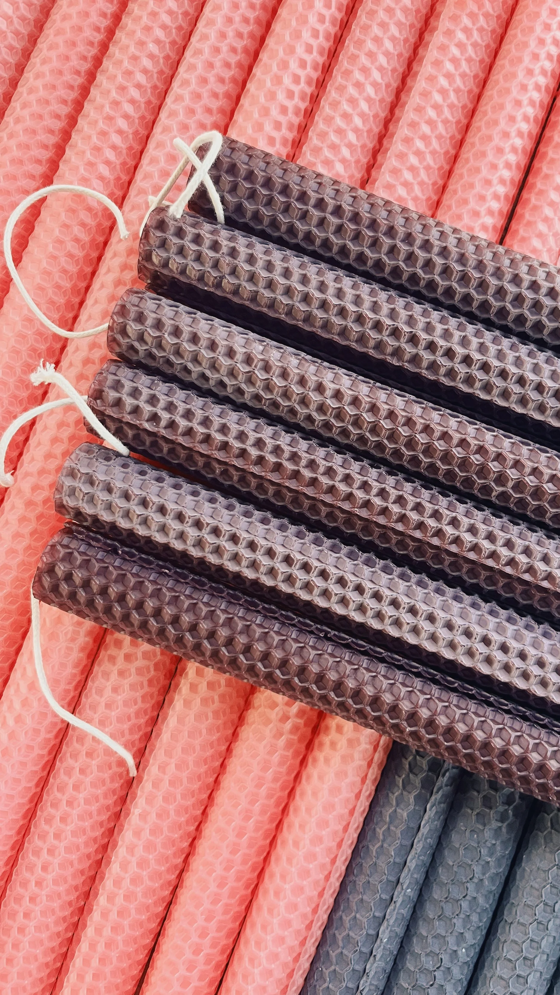 A stack of deep purple hand-rolled honeycomb beeswax taper candles with cotton wicks laid diagonally across rows of soft pink and black beeswax tapers.