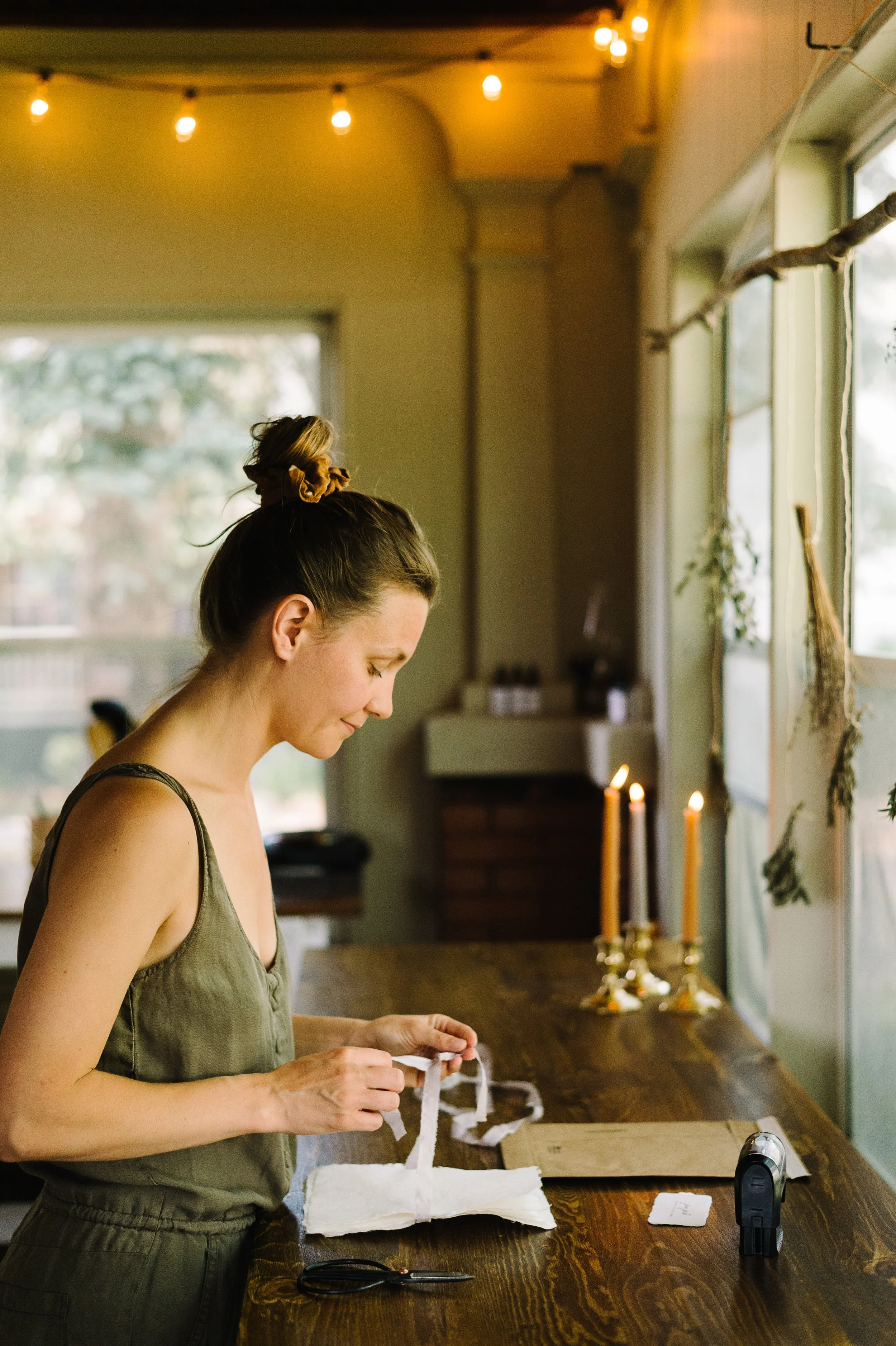 A woman with a bun hairstyle is tying silk ribbon around handmade paper at a wooden table. There are lit candles and a large window with trees outside in a cozy room with string lights on the ceiling.