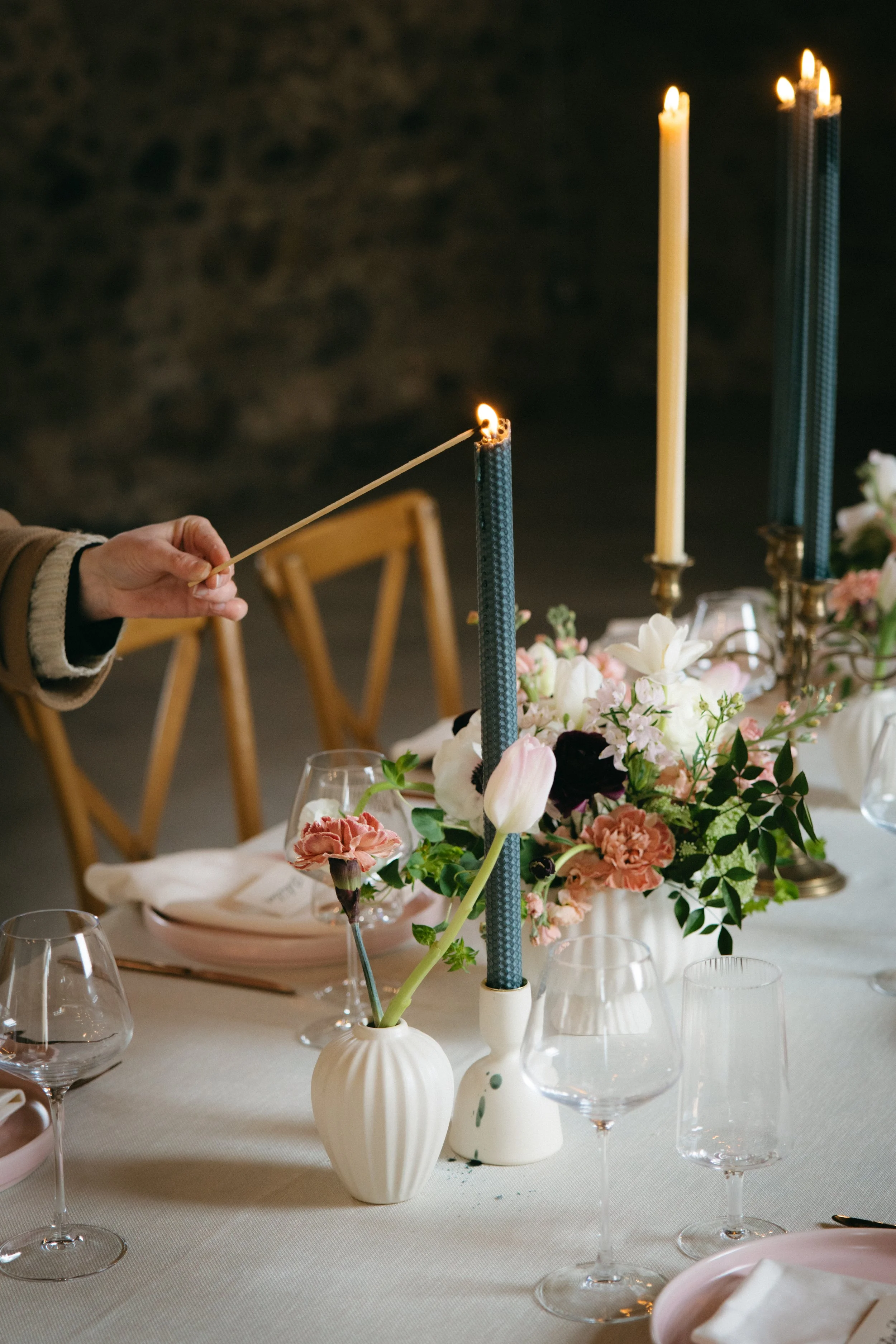 A person lighting a candle on a decorated table with floral centerpieces, wine glasses, and tableware, in a dimly lit setting.