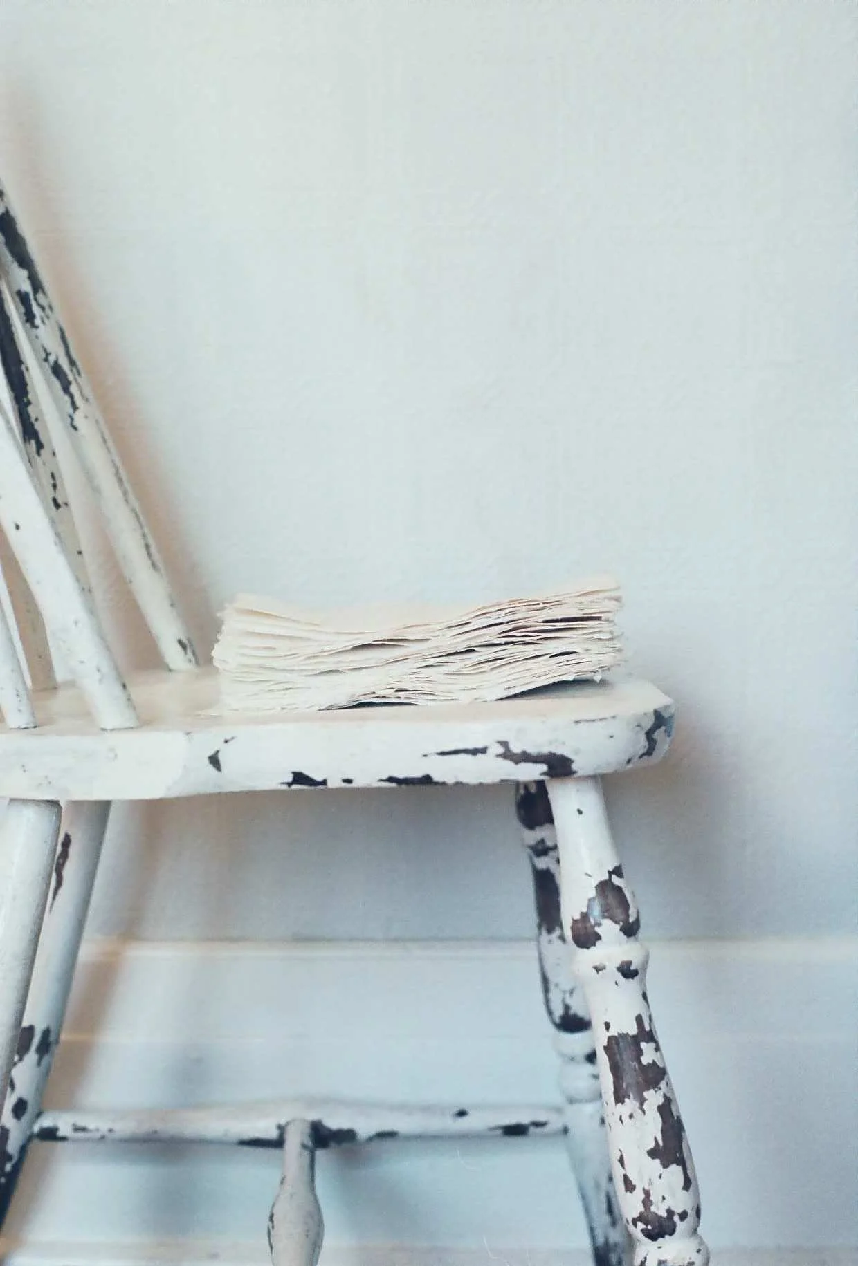 A vintage, rustic wooden chair with worn white paint holding a stack of textured handmade papers on its seat.