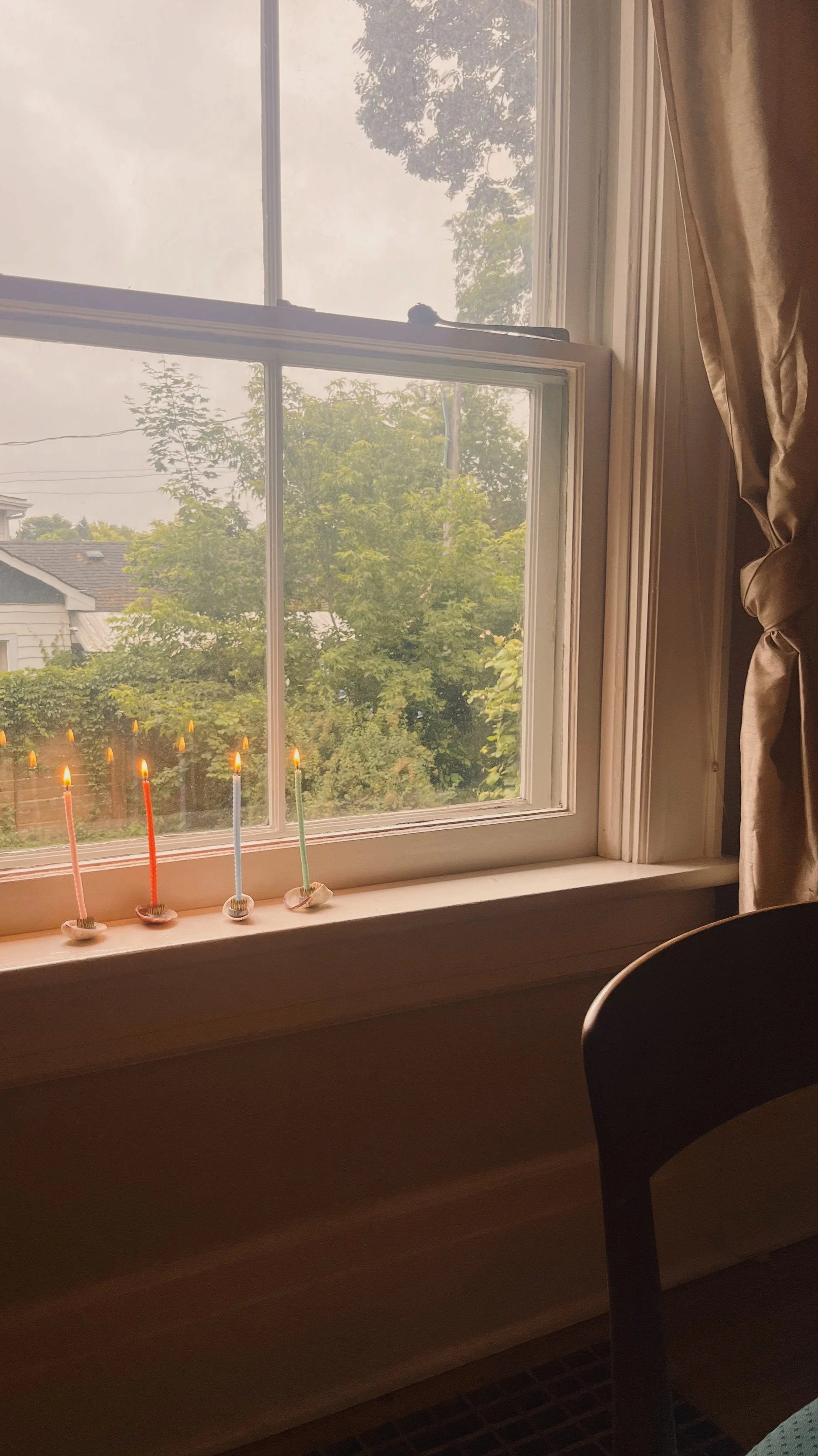 Four slender, colorful taper candles burning on a windowsill in small shell holders, with framed beige curtains and green trees visible through the glass outside