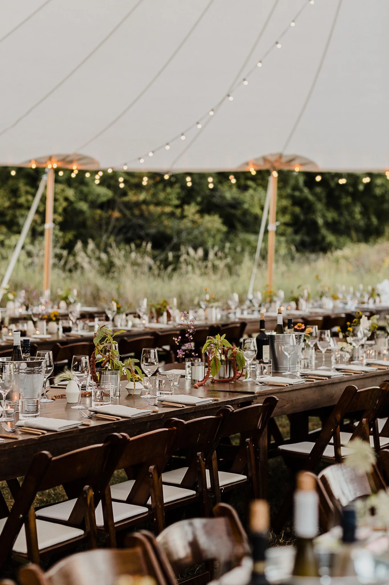 Outdoor wedding reception setup with long wooden tables, white napkins, wine bottles, glasses, and floral centerpieces under a large white canopy decorated with string lights, with trees in the background.