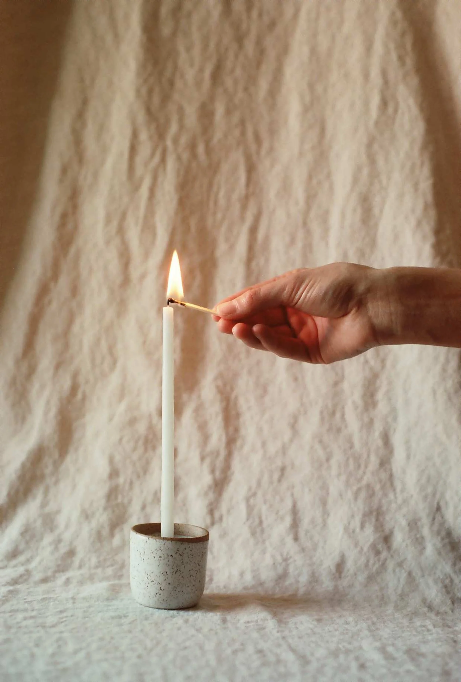 Hand lighting a tall white candle with a match in a small ceramic holder on linen at studio free folk.