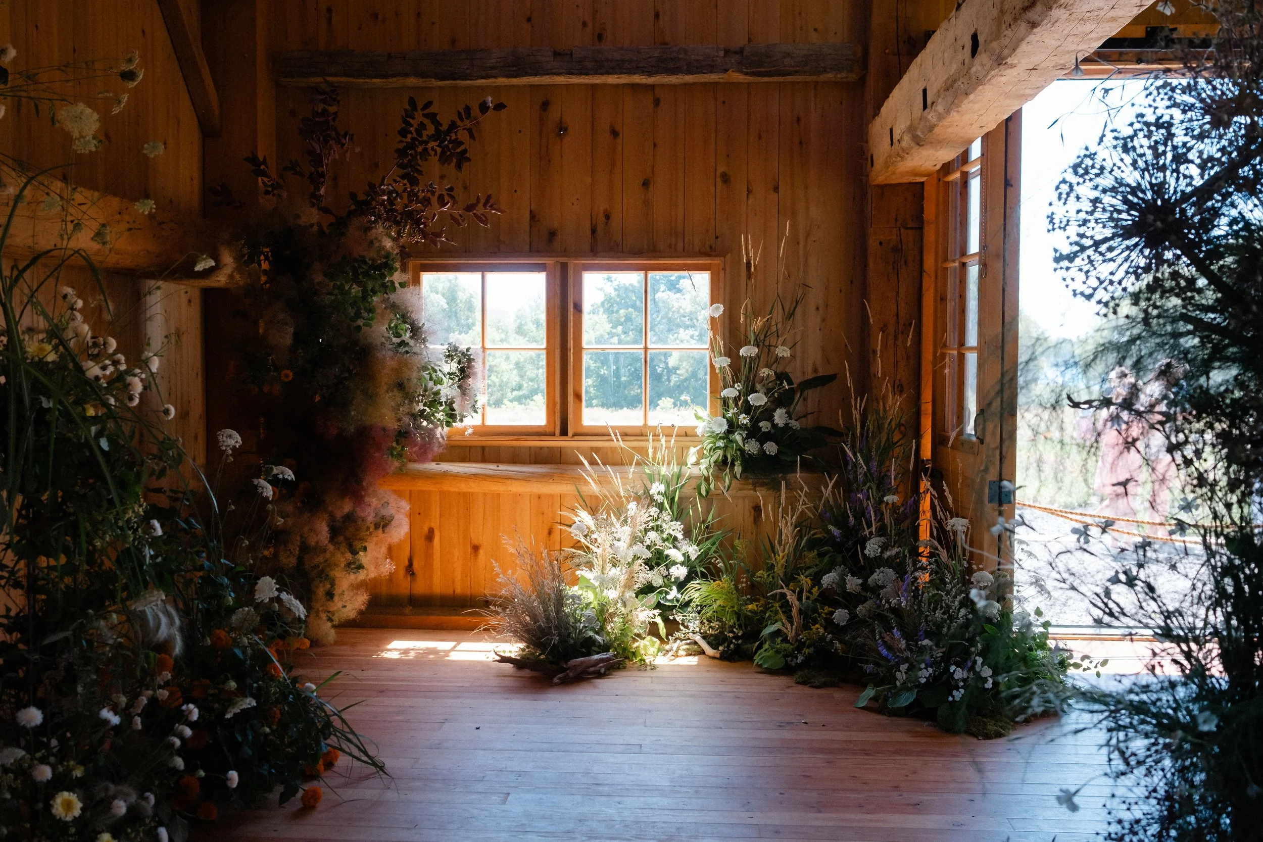 Interior of a rustic wooden barn decorated with a variety of flowers and plants for Flowering Wild, with a large window letting in natural light.