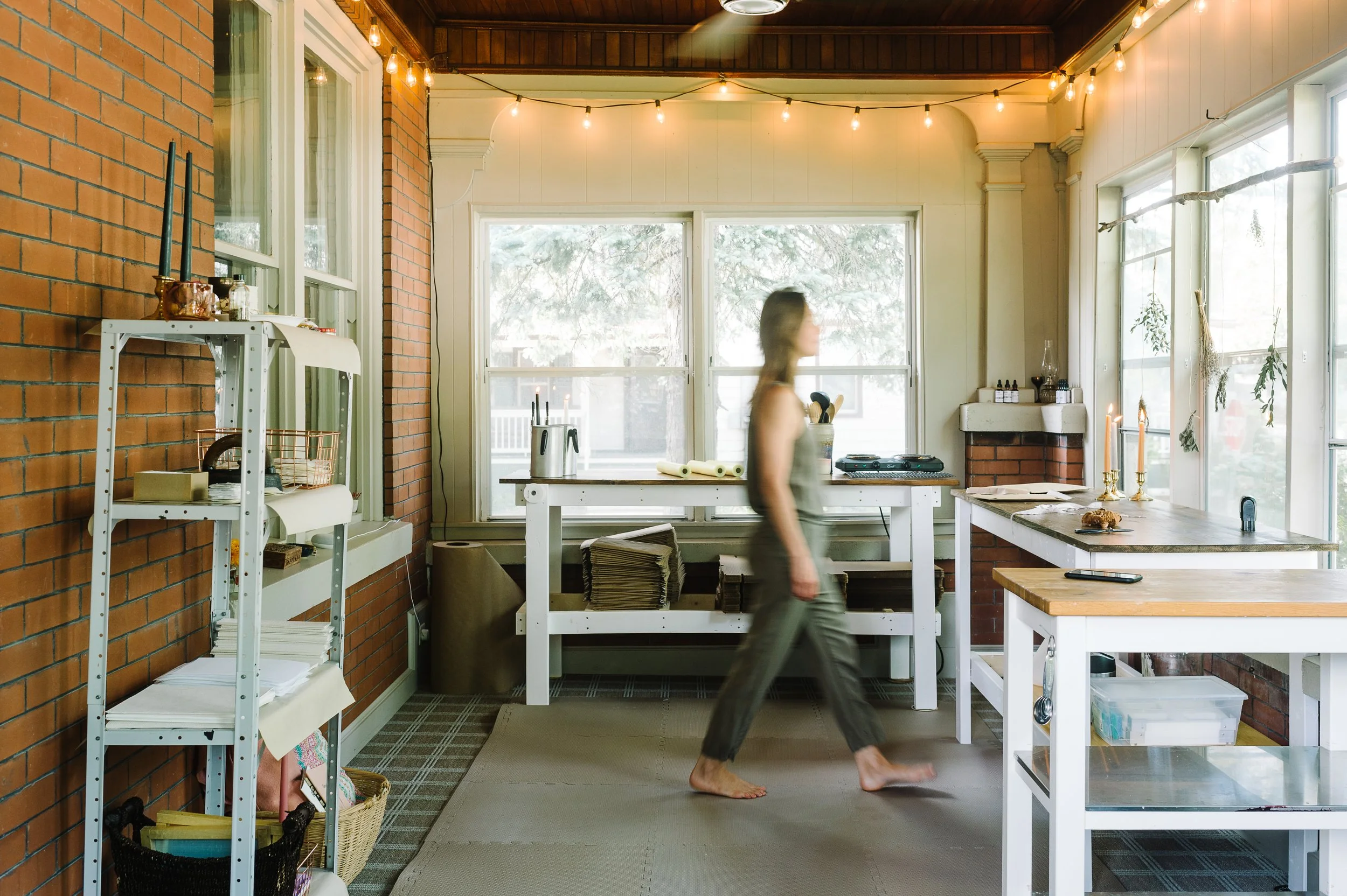 A woman walking barefoot through a bright studio with large windows, string lights, brick walls, and various candle making supplies found objects, and art on shelves and counters.