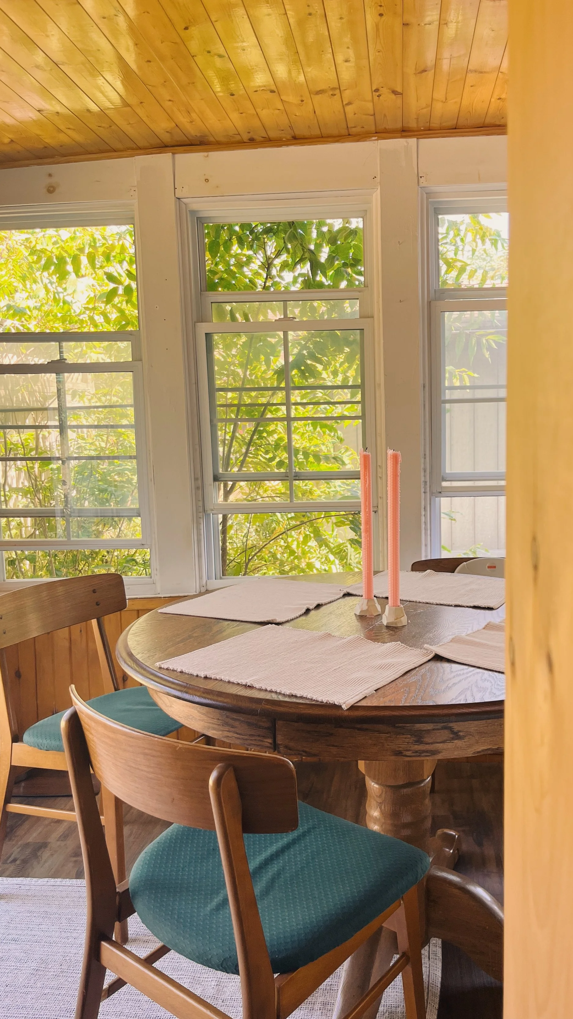 A pair of tall pink hand-rolled beeswax taper candles set on a round wooden dining table in a bright, sunlit room with large windows and lush green trees outside.