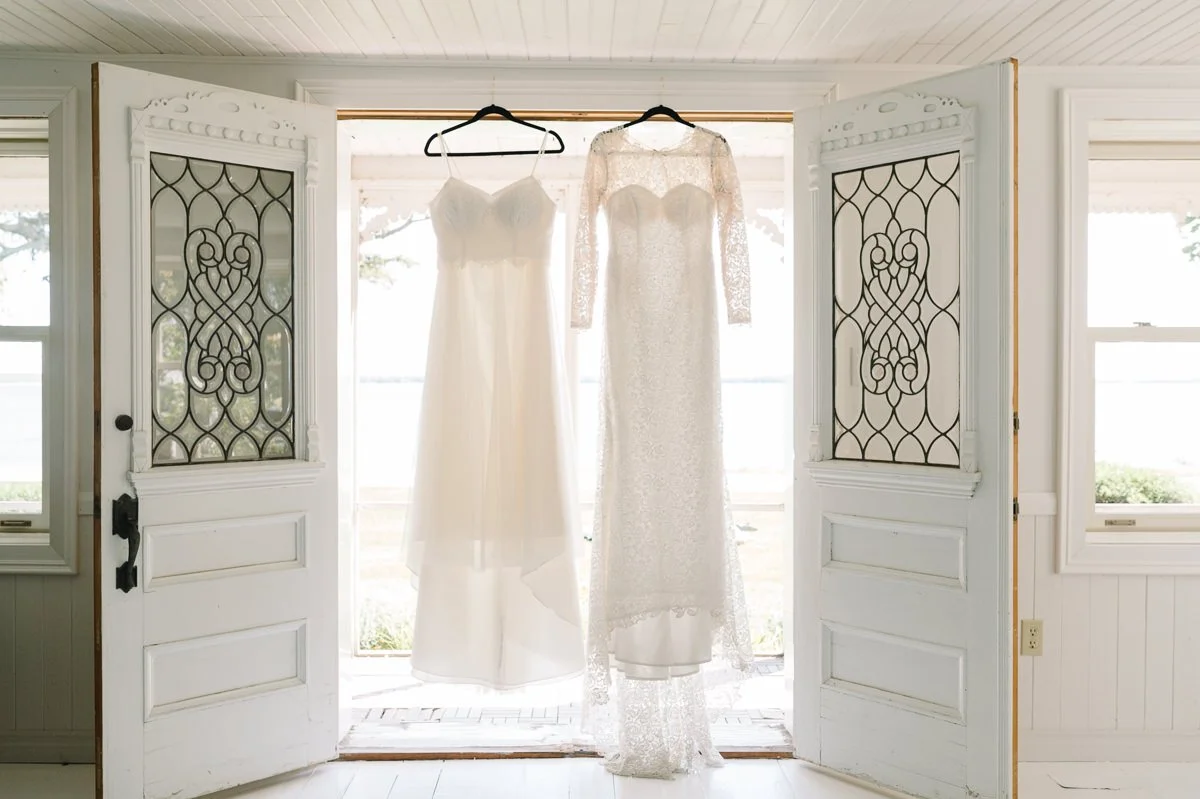 Two wedding dresses hanging inside a porch area in front of two open doors with decorative glass panels.