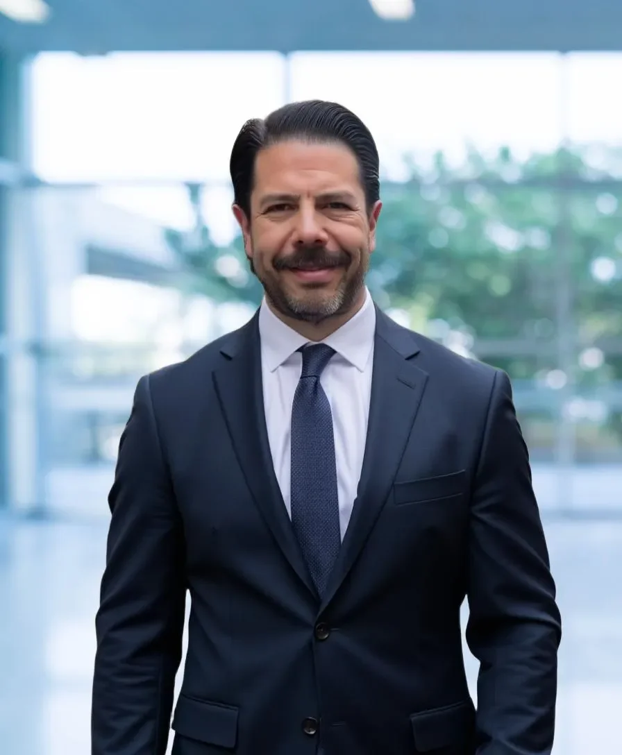 A middle-aged man with dark hair, a beard, and a mustache, dressed in a dark suit, white shirt, and blue tie, standing indoors with a blurred background of large windows and greenery outside.