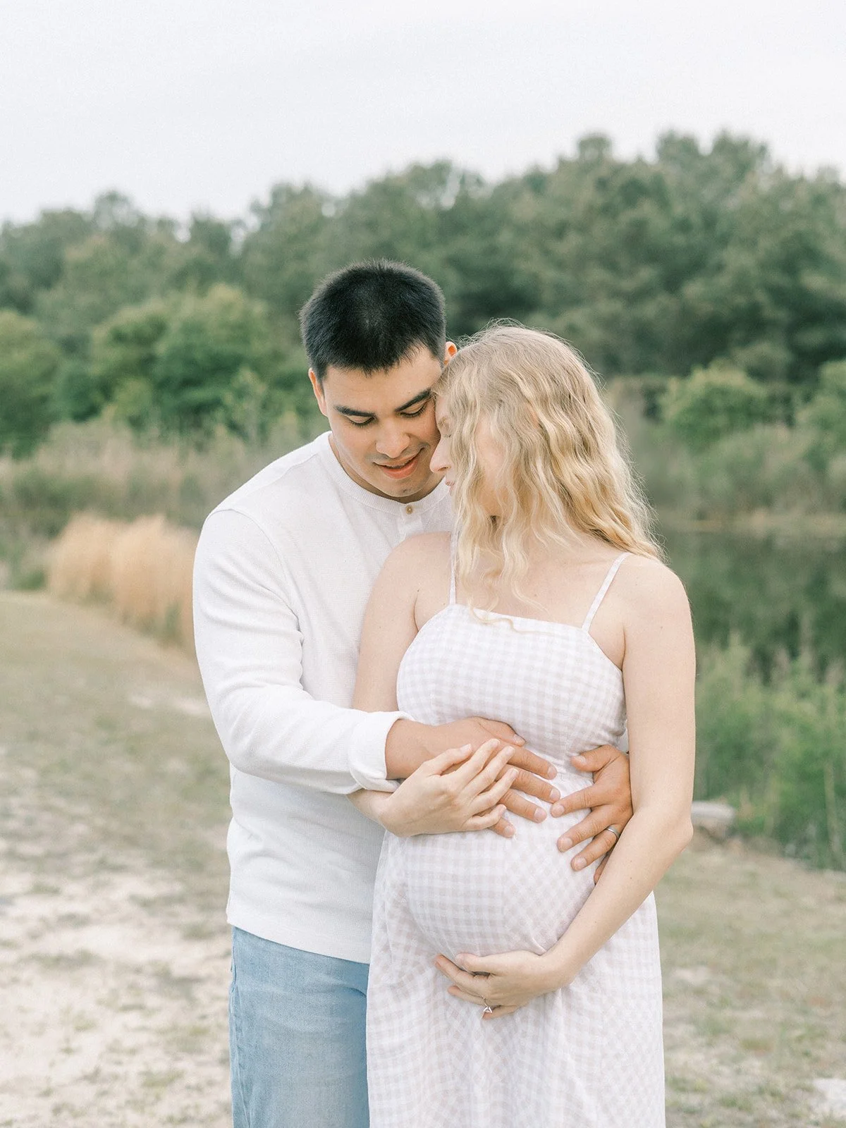 A pregnant woman and her partner standing outside near a body of water, embracing each other lovingly with their heads touching, in a natural setting with trees and grass in Virginia Beach.