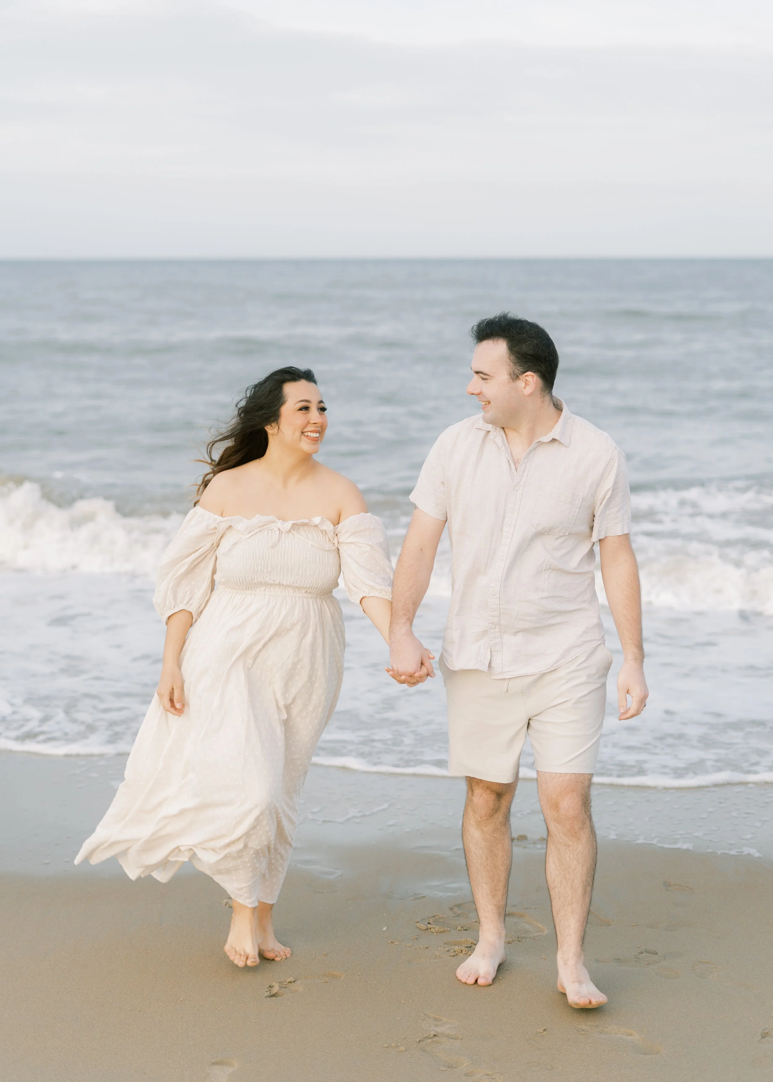 A smiling couple holding hands and walking on the beach near the water, dressed in light-colored casual clothes.