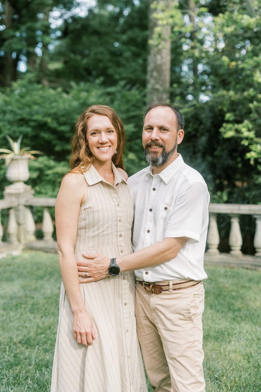A smiling man and woman standing outdoors on a green lawn with trees and a stone railing in the background.
