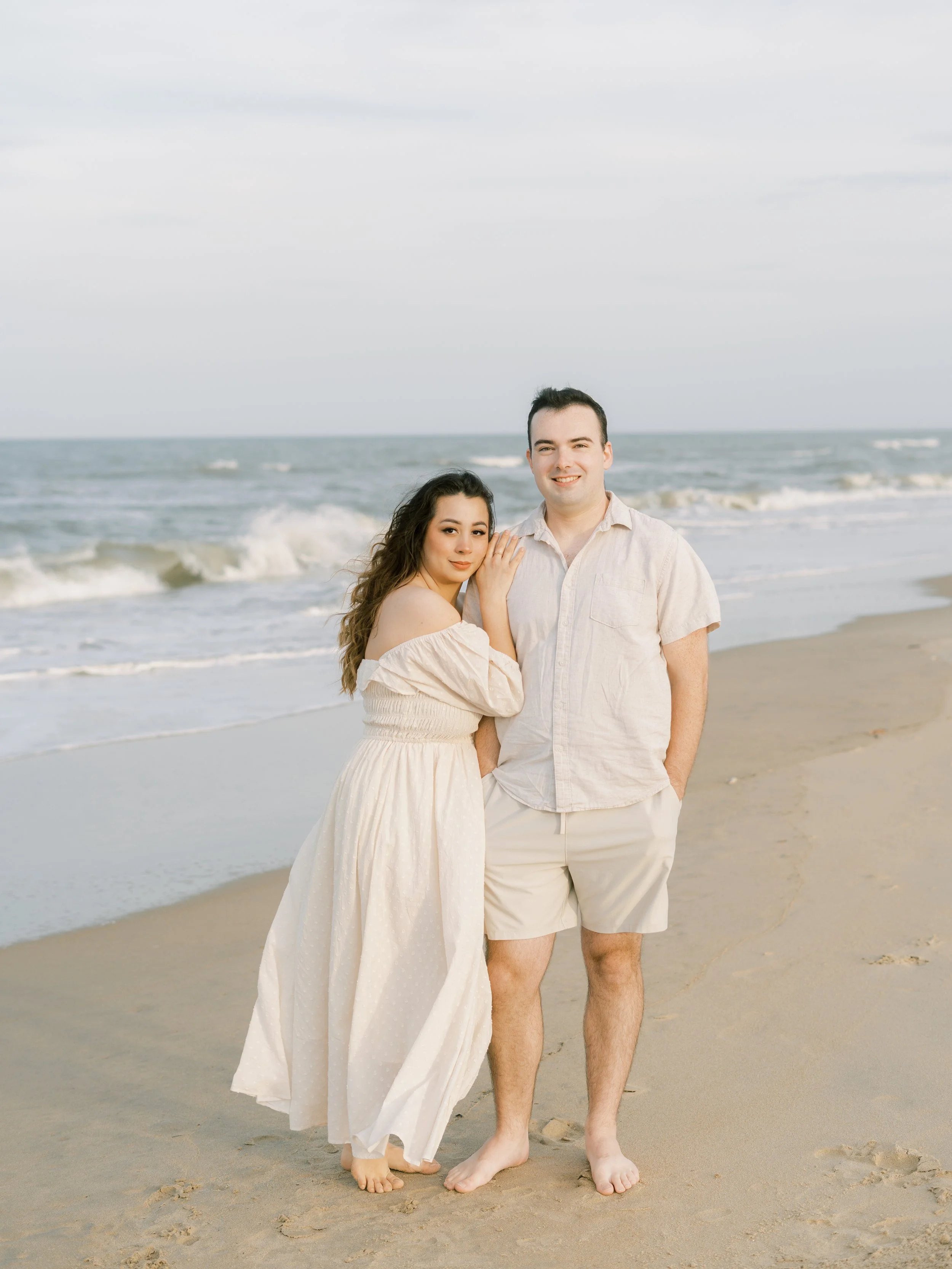 A young couple standing on the beach with the ocean and sky in the background, smiling and posing for a photo.