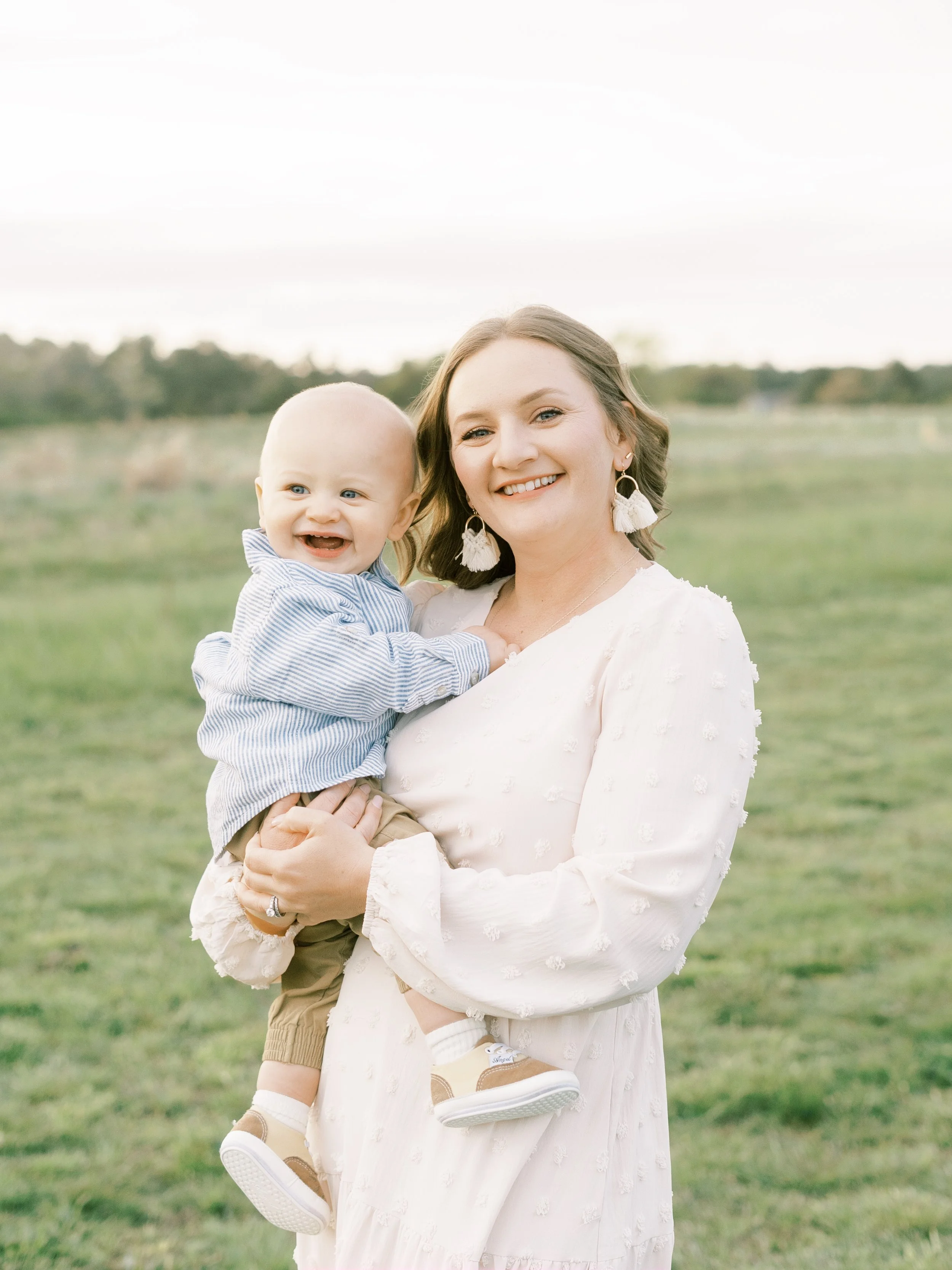 A woman holding a smiling baby boy outdoors in a grassy field with a blurred tree line in the background.