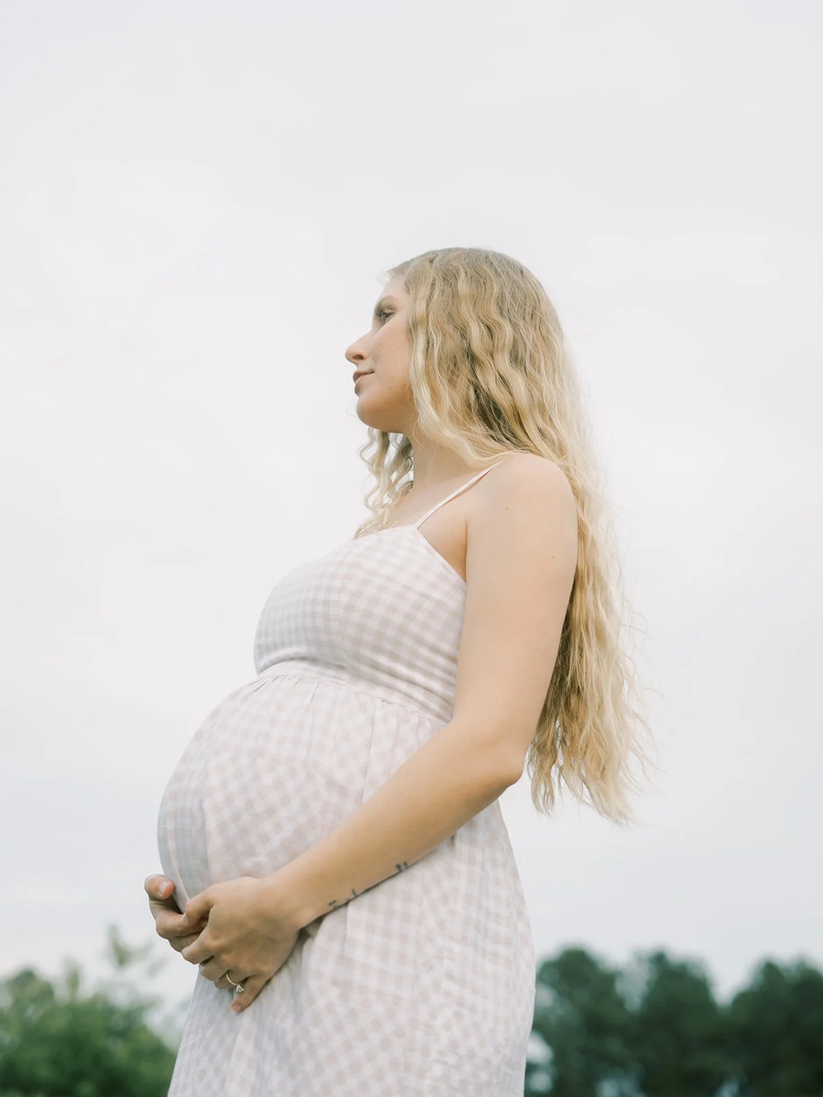 Pregnant woman with long wavy blonde hair standing outdoors against a cloudy sky, wearing a sleeveless, light-colored, checkered dress.