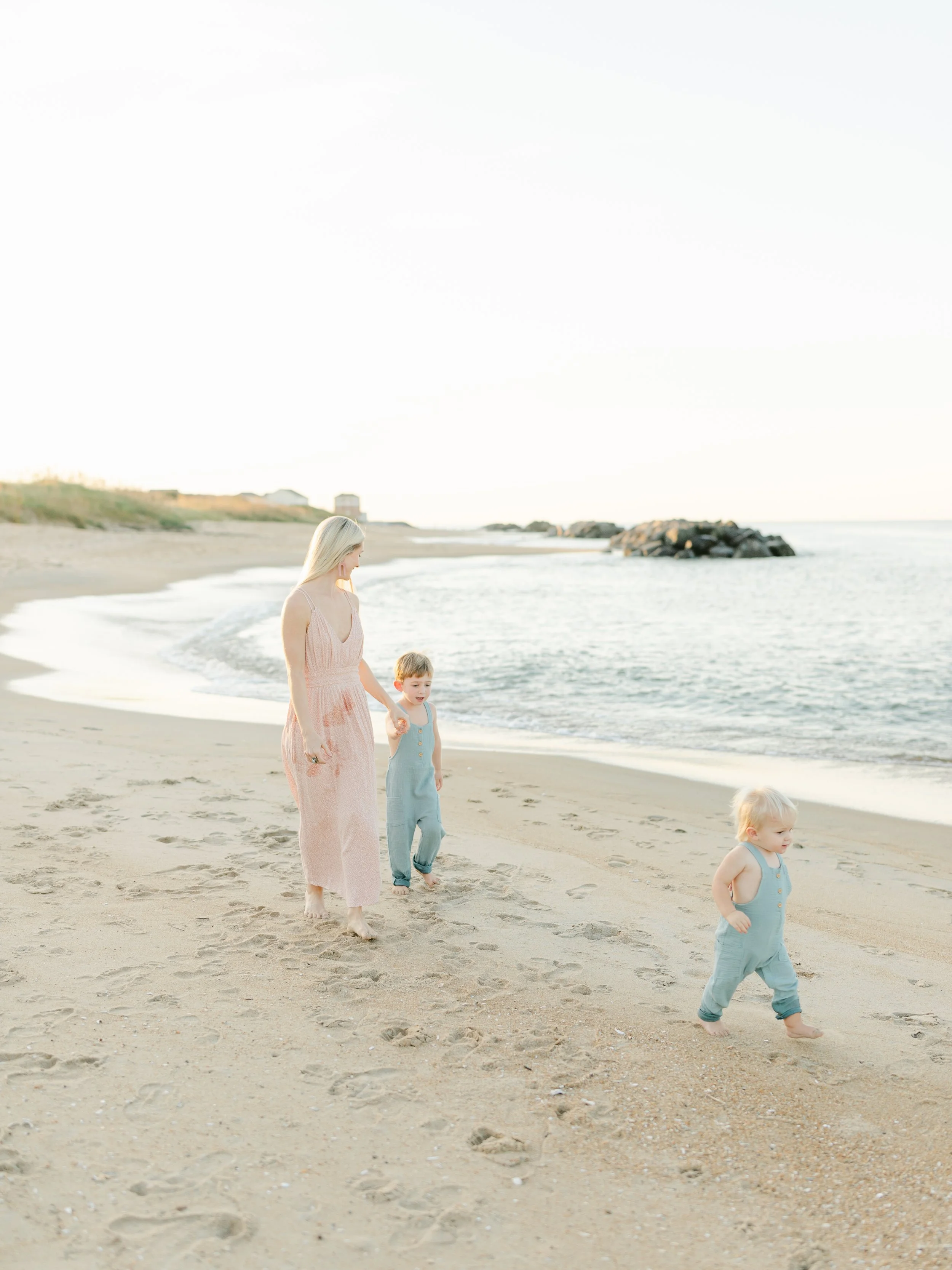 A woman in a pink dress walks along the beach with two small children in blue outfits. The ocean is visible in the background, with rocks on the shoreline and a clear sky.