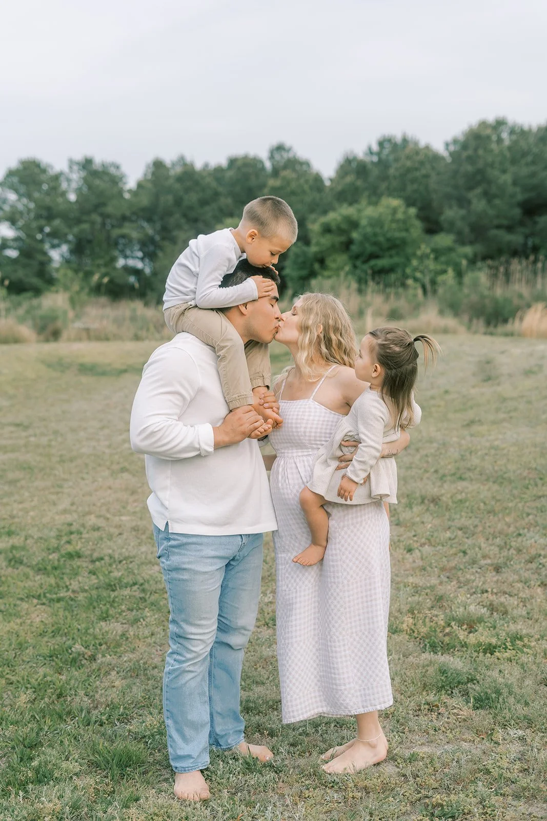 A family of five outdoors on a grassy field, with a man holding a boy on his shoulders, and a woman holding a girl, as they share a kiss.