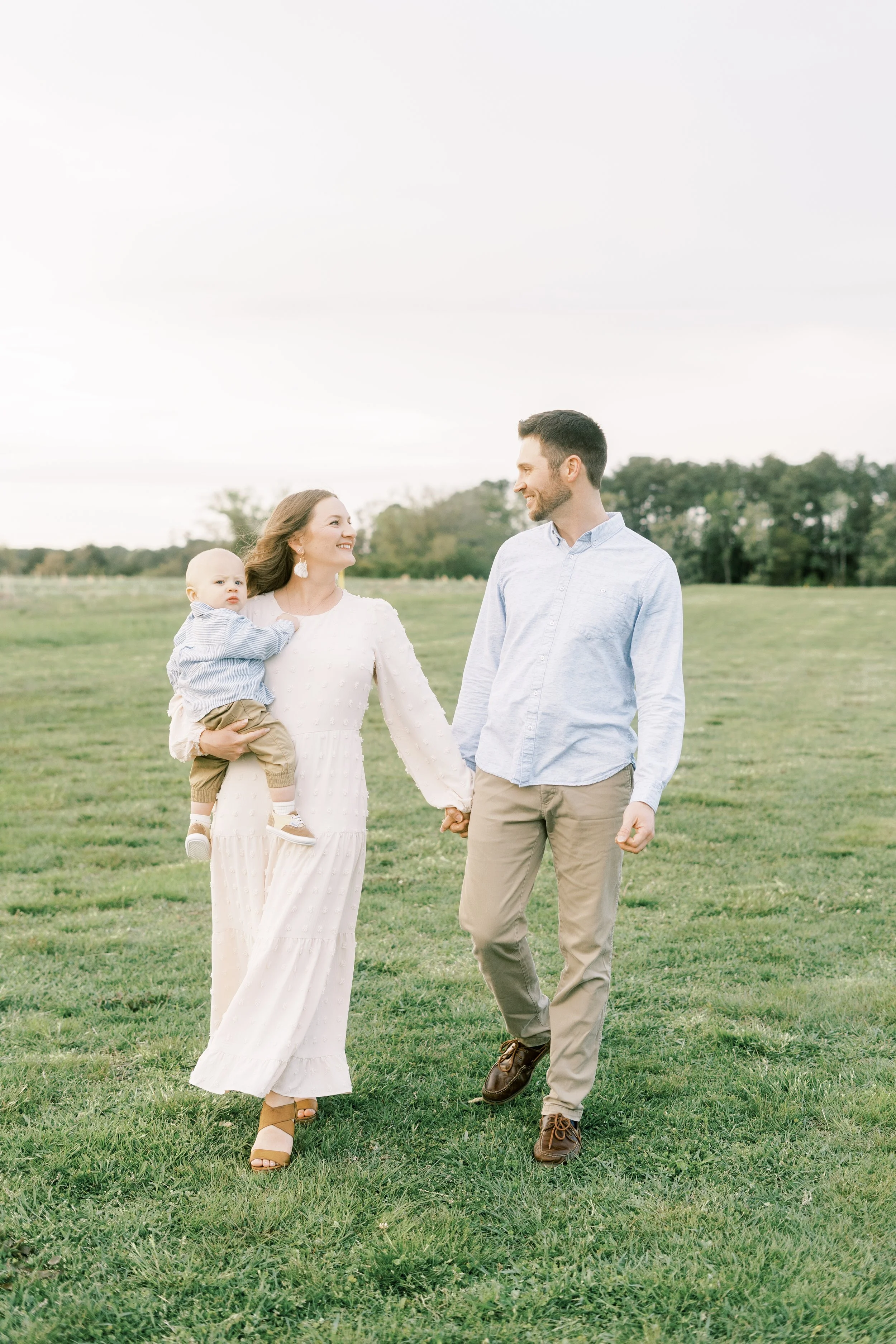A happy family walking hand in hand on a grassy field, with a woman holding a toddler, and a man walking beside them, all smiling at each other.