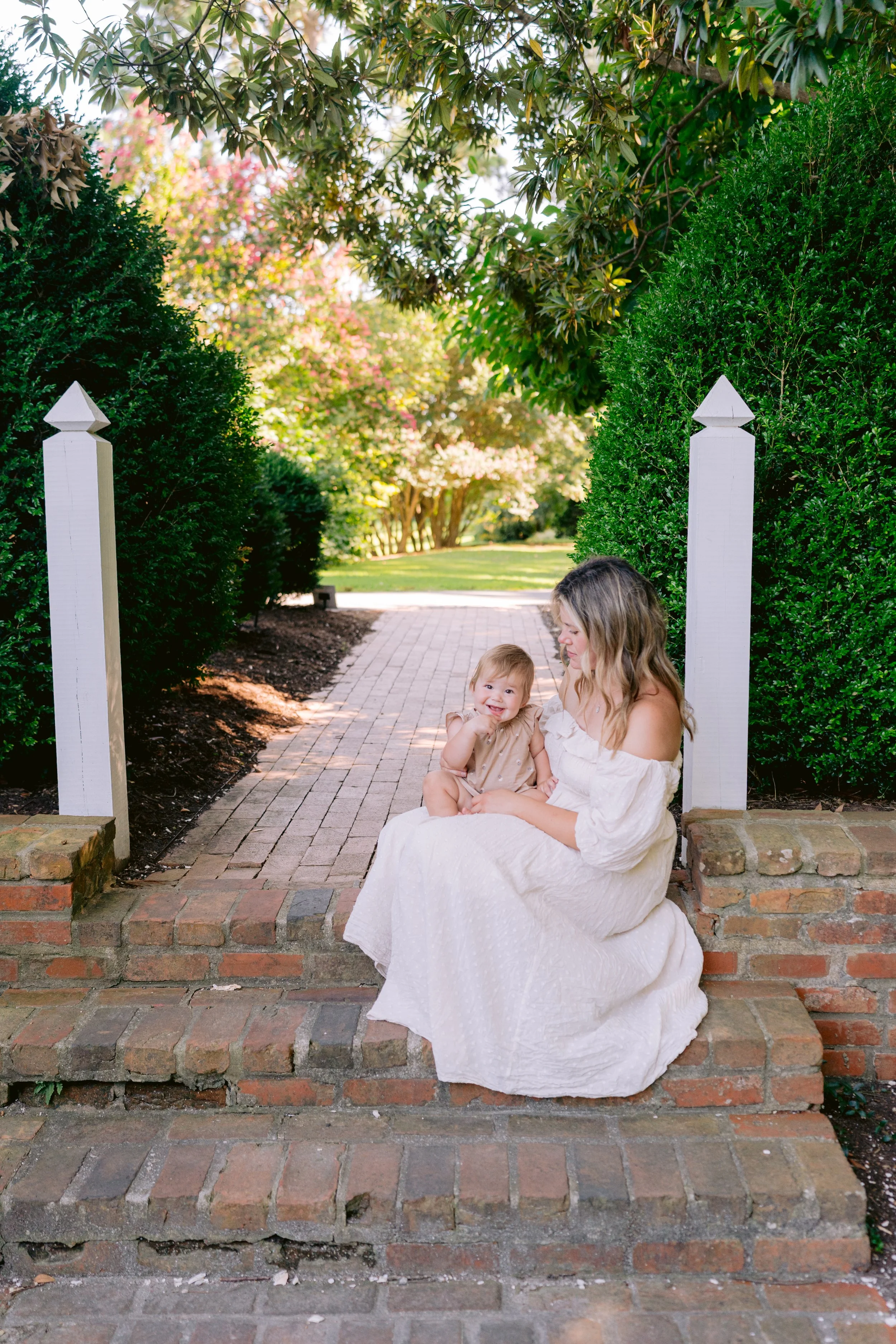 A woman in a white dress sitting on brick steps outdoors, holding a smiling toddler in a beige outfit, with green trees and a sidewalk in the background.