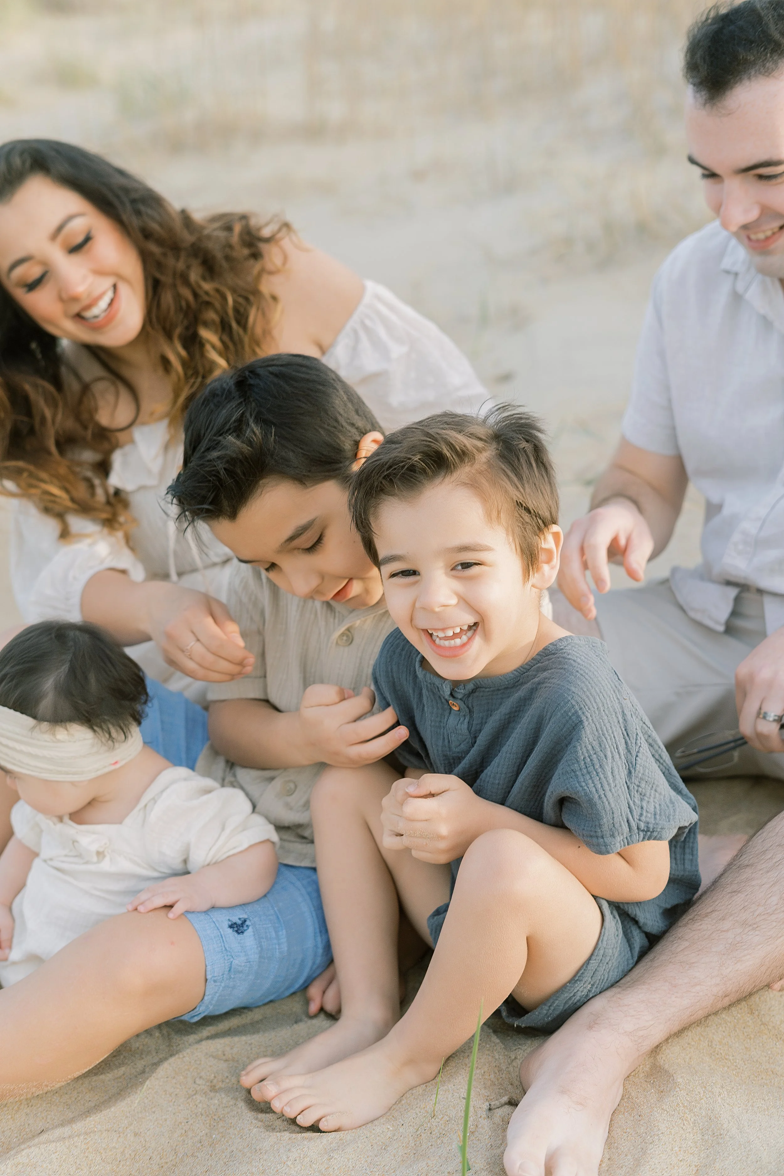 A happy family of six, including two young children and two babies, sitting on the sand outdoors, smiling and interacting with each other.
