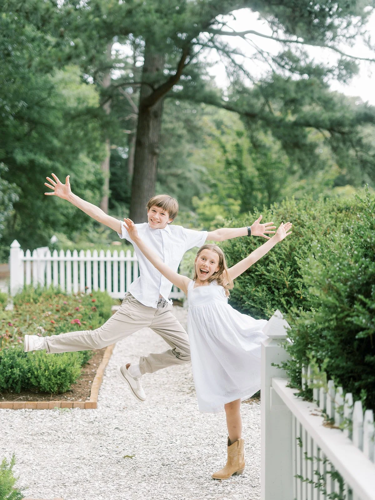 Two children, a boy and a girl, are jumping with arms outstretched on a gravel path in a lush green garden, smiling happily.