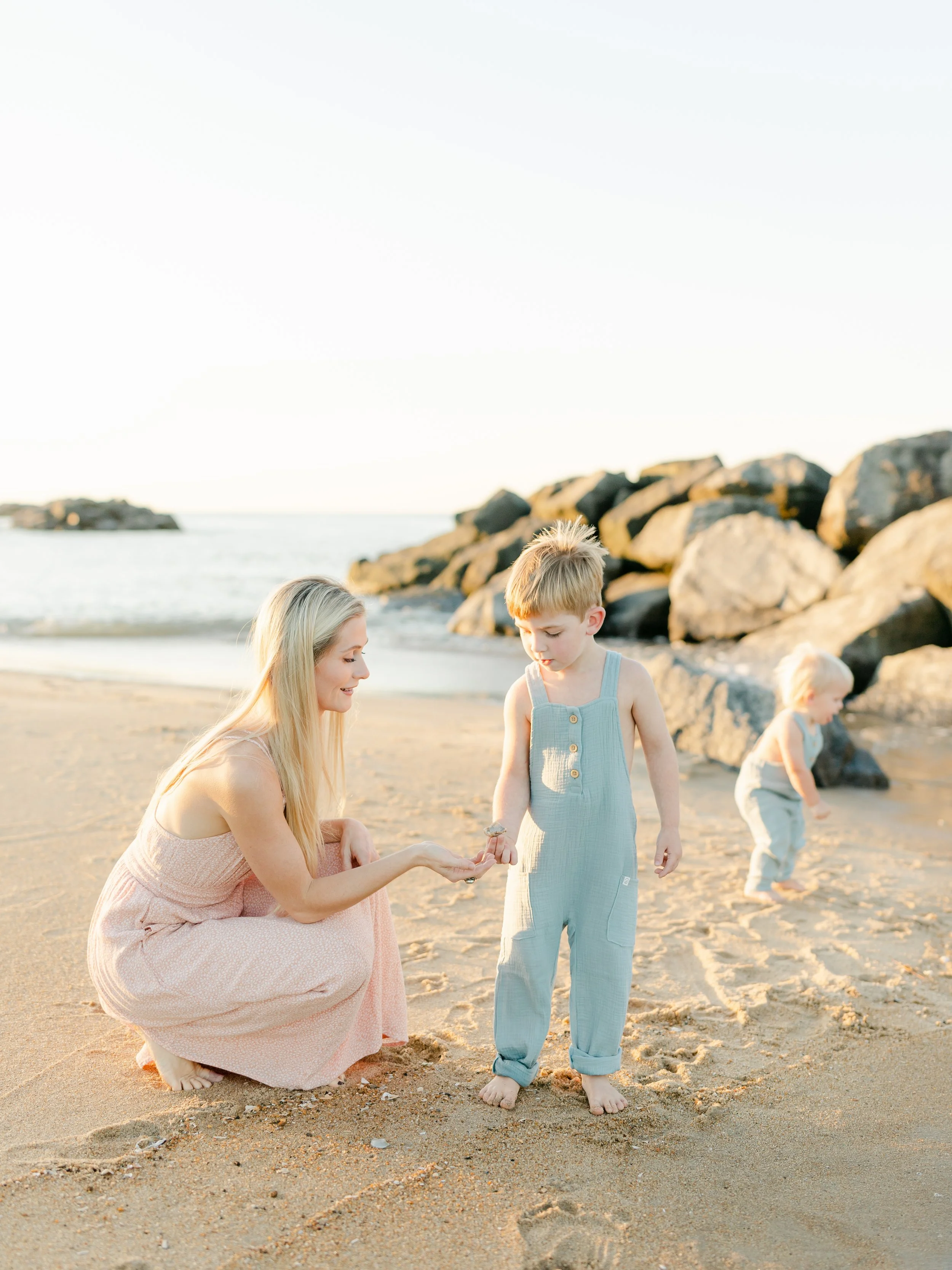 A woman kneeling on a sandy beach showing a shell to a young child in blue overalls. Another child is walking in the background near some rocks. The scene is sunlit, and the ocean is visible.