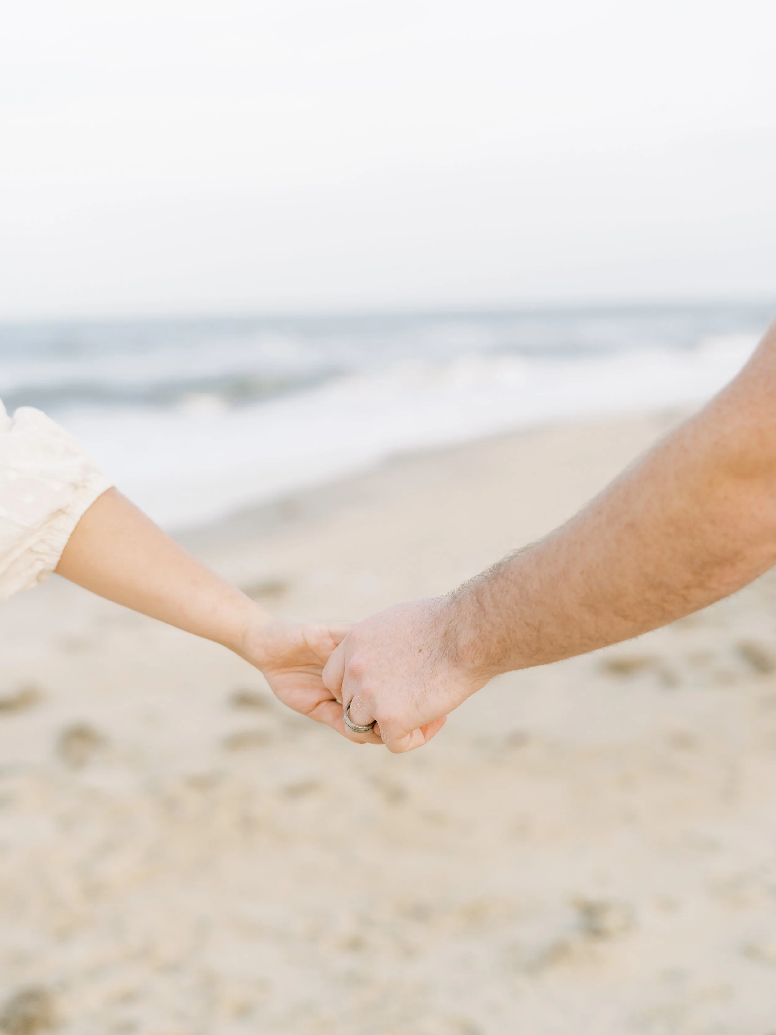 A man and woman holding hands on a sandy beach with the ocean in the background.