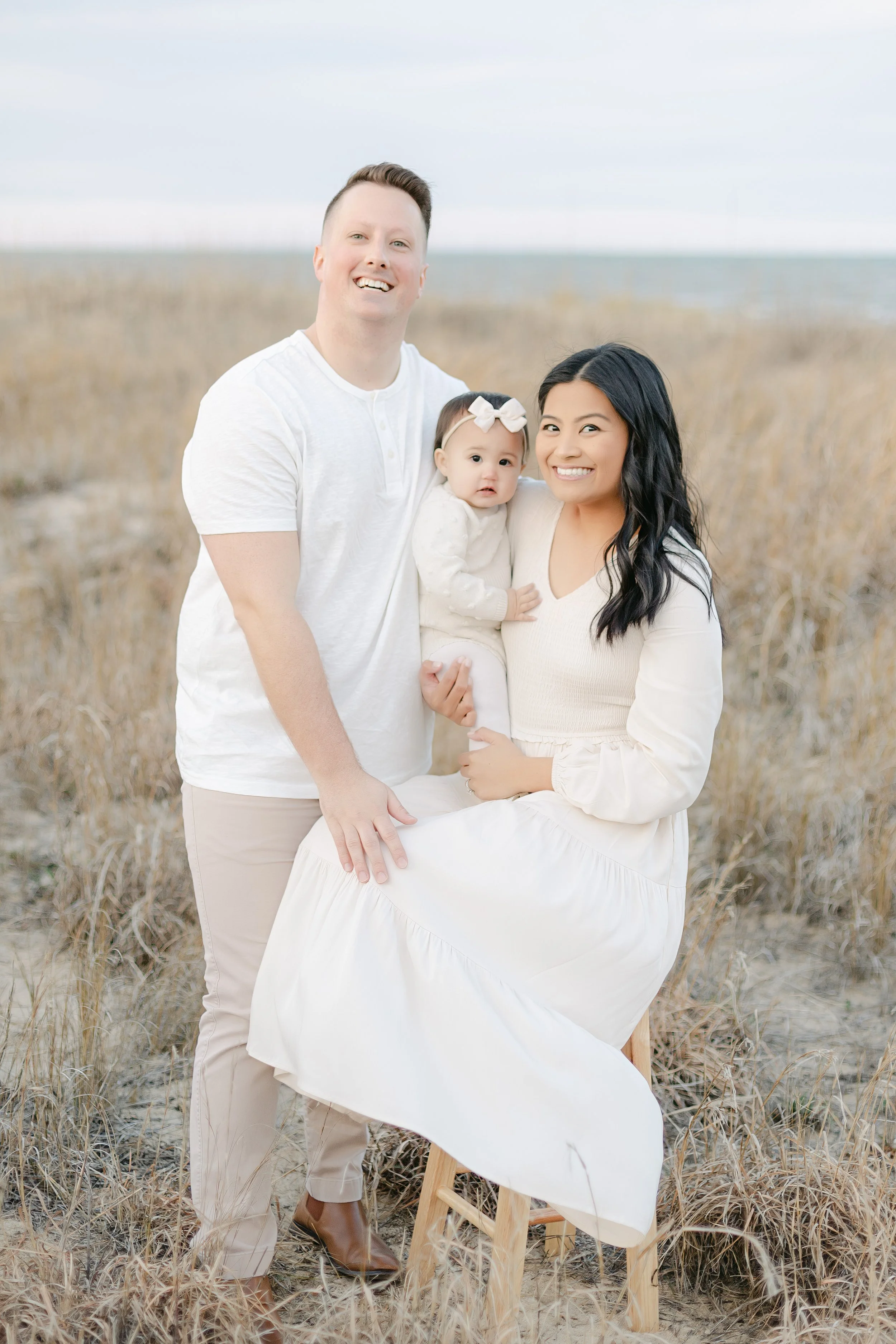 A family of three posing outdoors in a grassy field, with a man in a white shirt, a woman in a white dress holding a baby with a bow, all smiling.