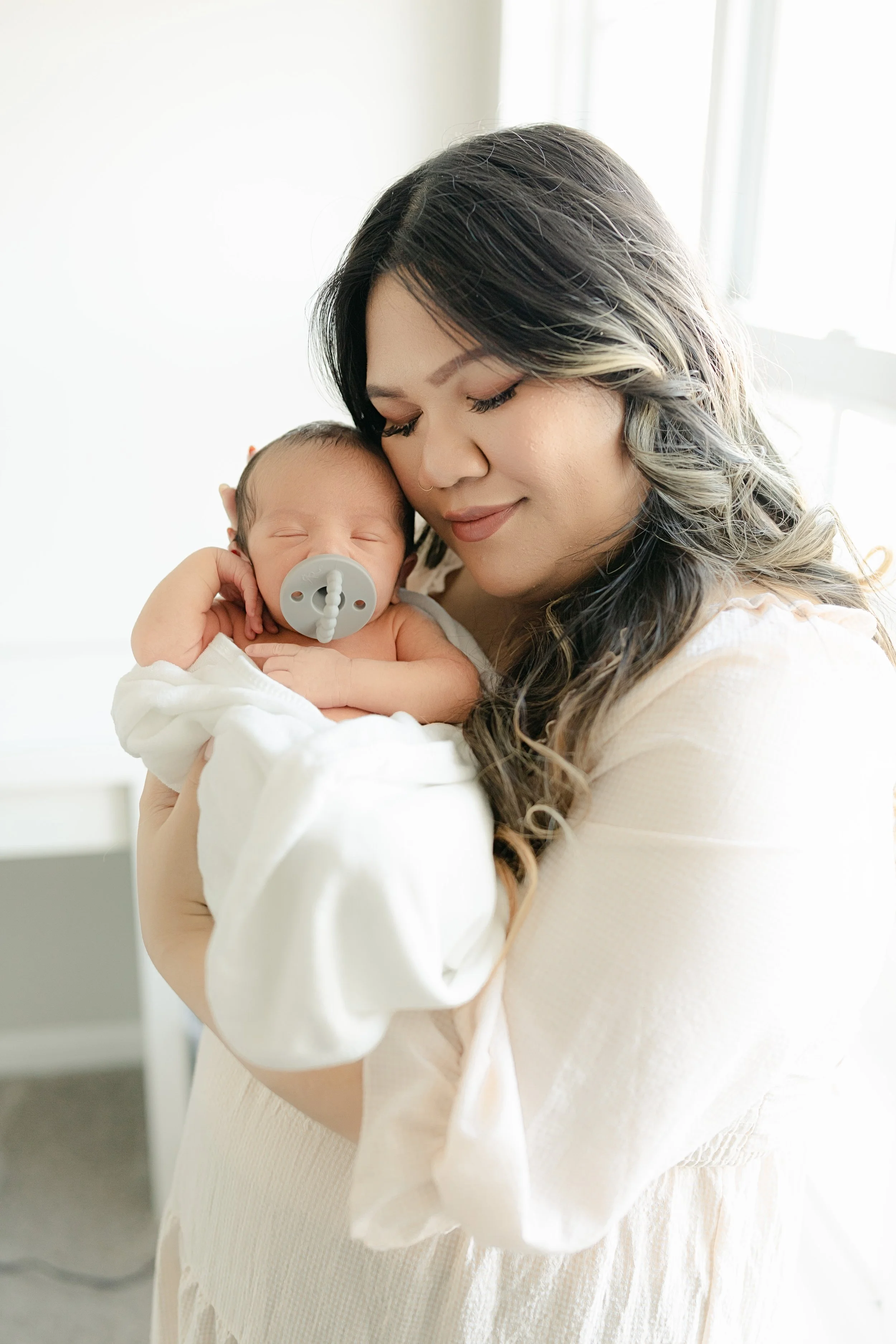 Woman holding a sleeping newborn wrapped in a white blanket with a pacifier in a softly lit room.