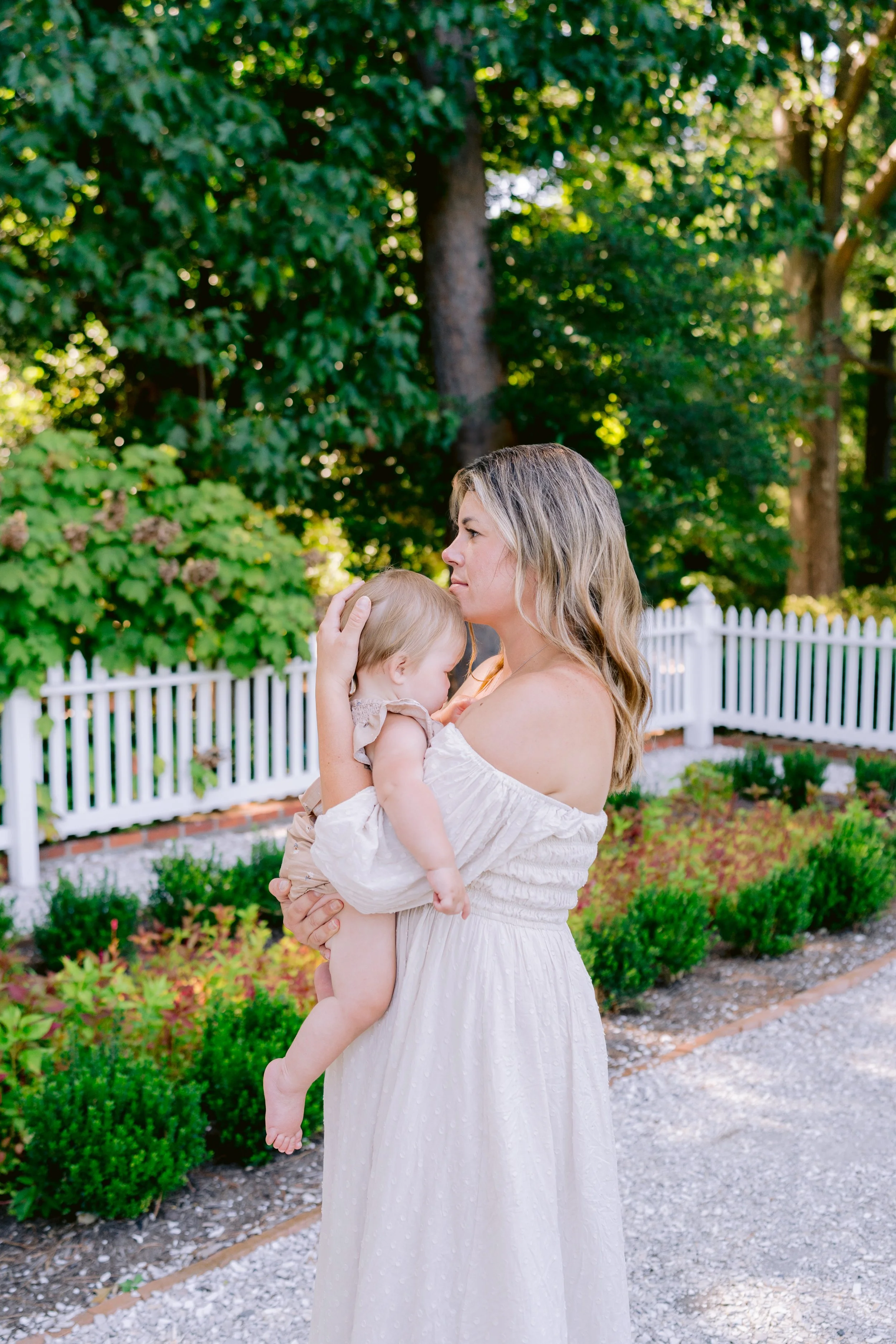 A woman with blonde hair wearing a white off-shoulder dress is holding a young child with blonde hair, also dressed in light colored clothing, in a garden with a white picket fence and green trees in the background.