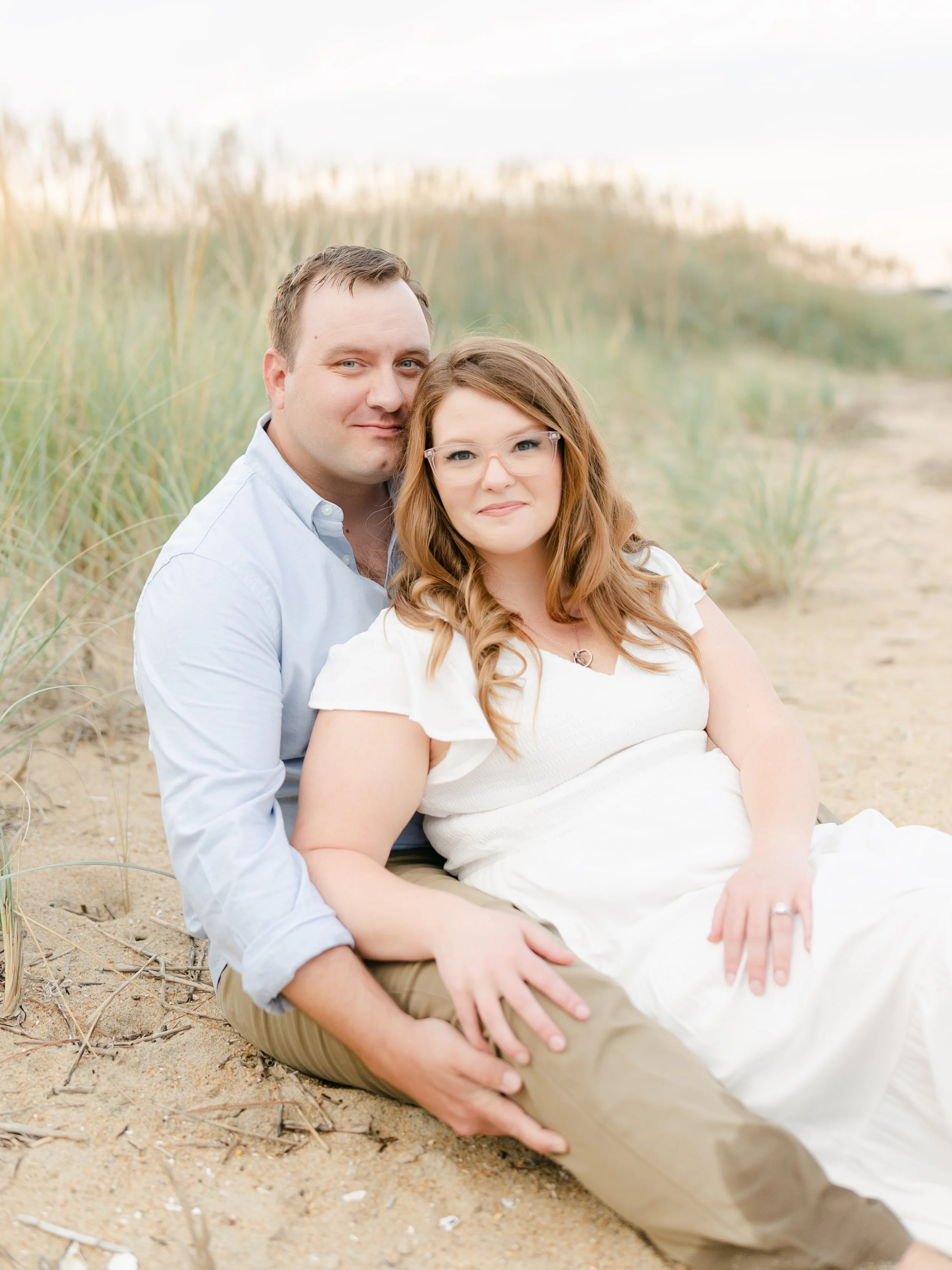 A couple sitting on a sandy beach with tall grasses in the background, dressed in light-colored clothing, with the woman wearing glasses and both looking at the camera.