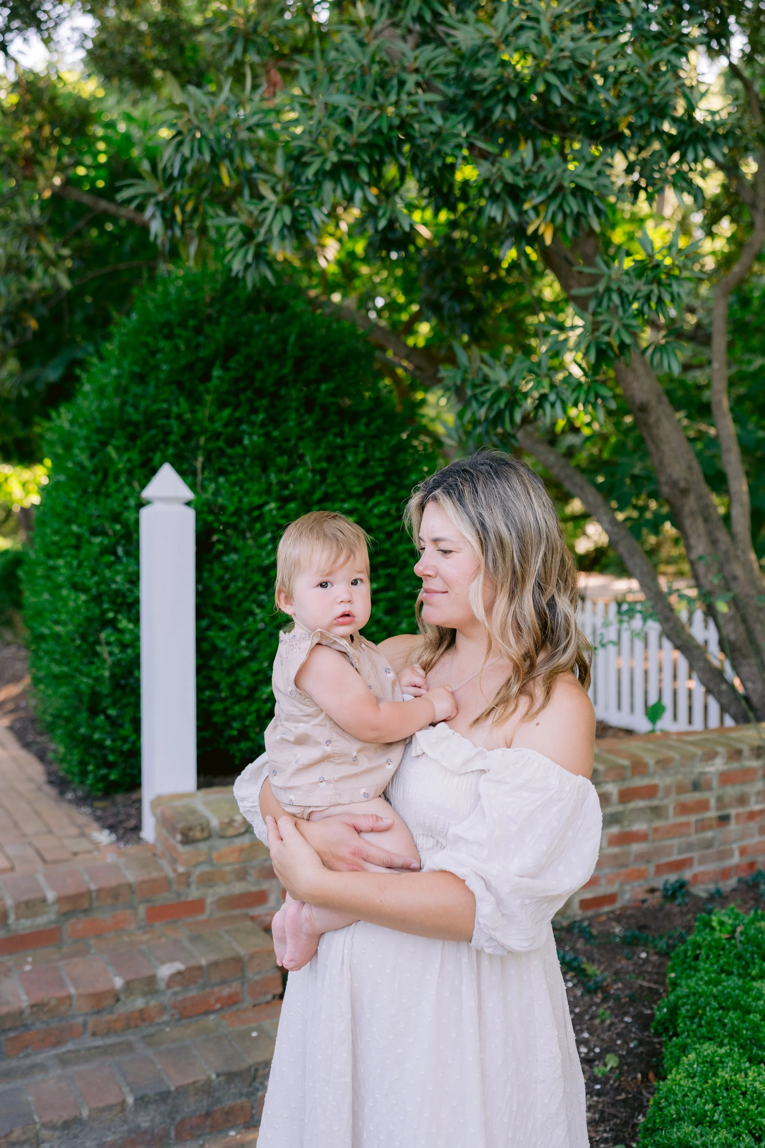 A woman with wavy blonde hair holding a young girl with light hair, standing outdoors in a garden with green bushes and trees, white picket fence, and brick path.