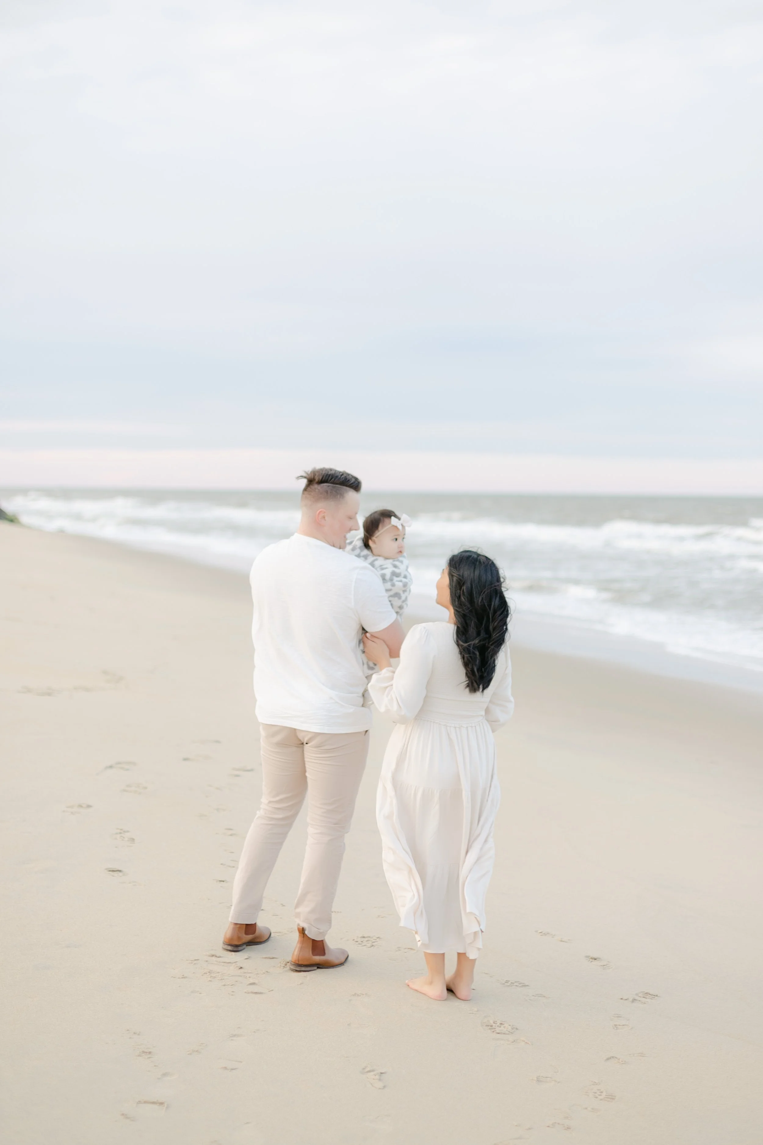 A family of three walking along a beach, with a man holding a child and a woman beside them. The ocean waves and sky are visible in the background.