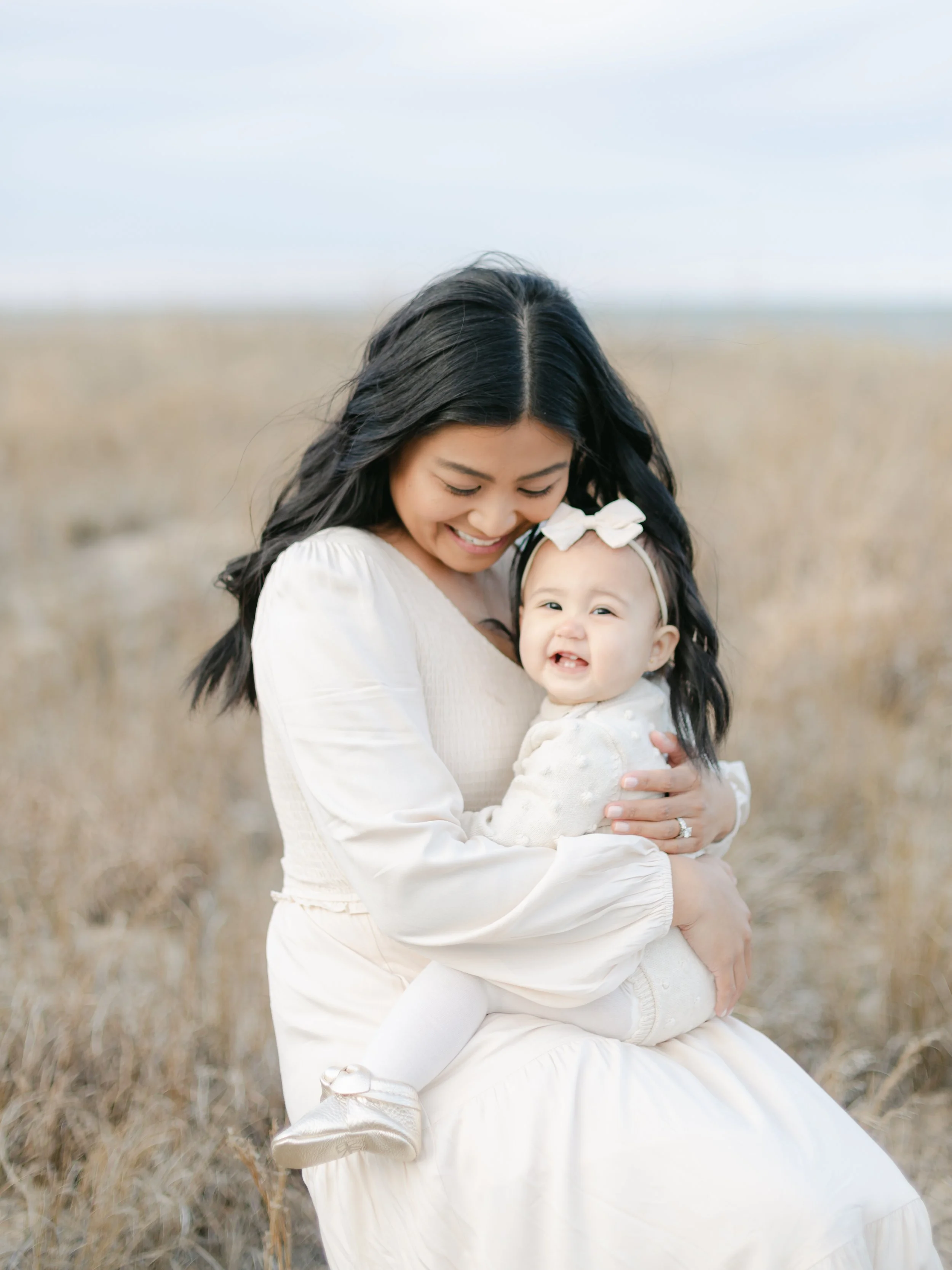 Woman holding a baby in a field, both wearing white clothes, with the baby smiling.
