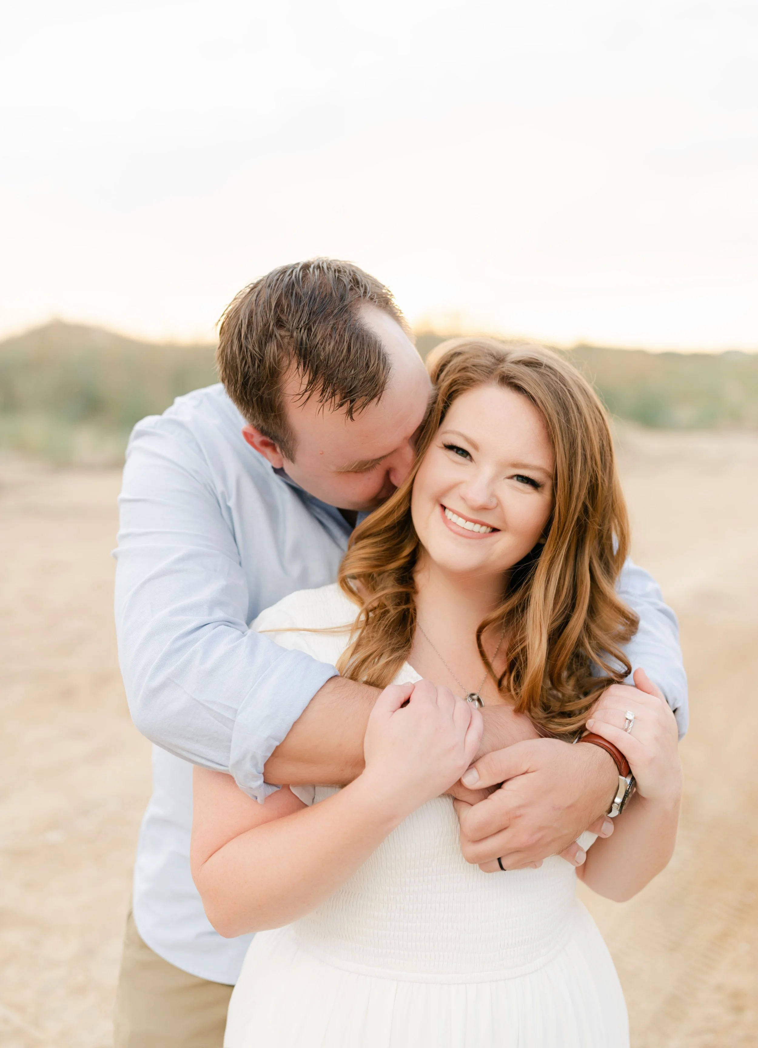 A young woman smiling while being embraced by a man from behind in an outdoor setting.