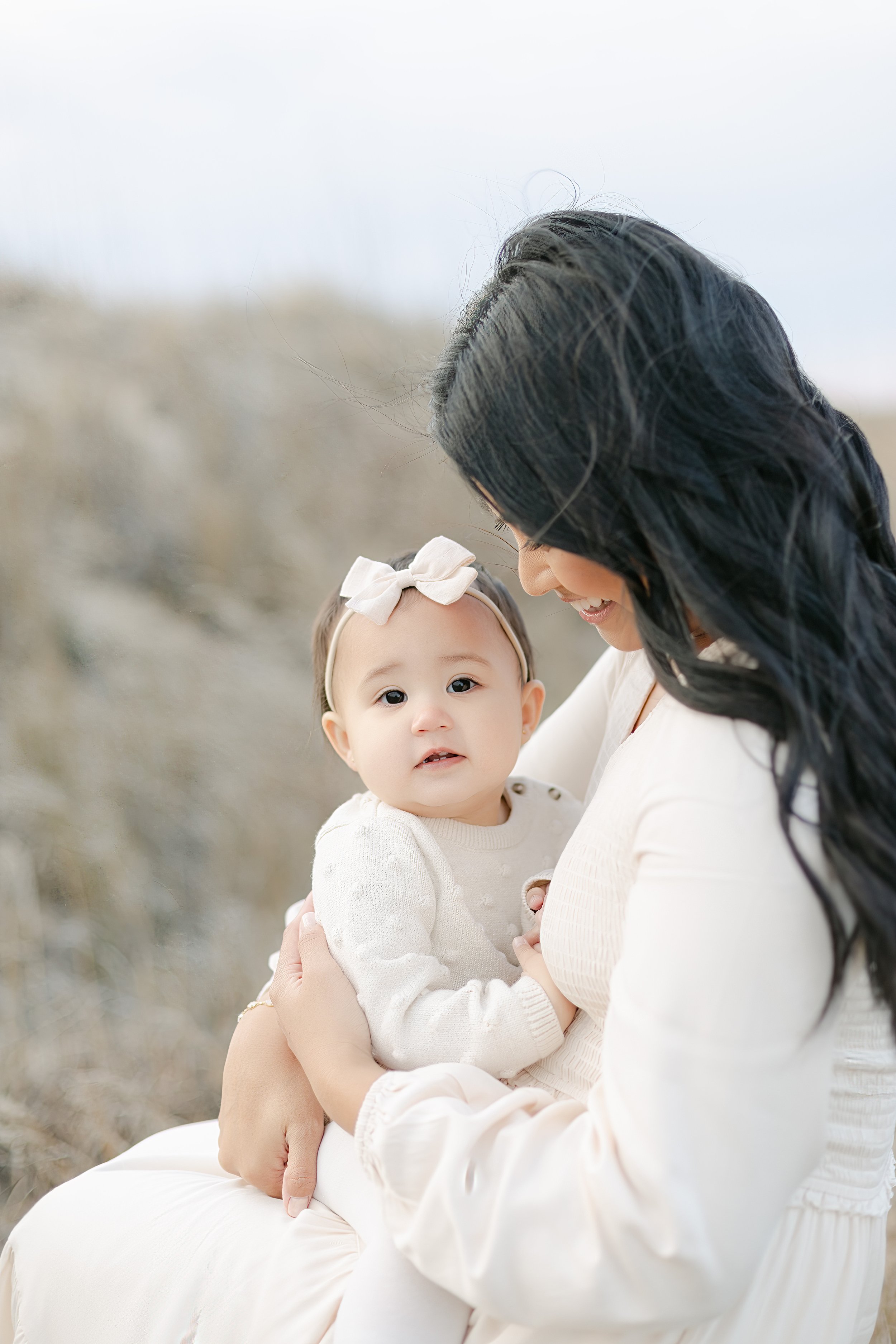 Woman holding a baby wearing a white outfit and bow in an outdoor setting.
