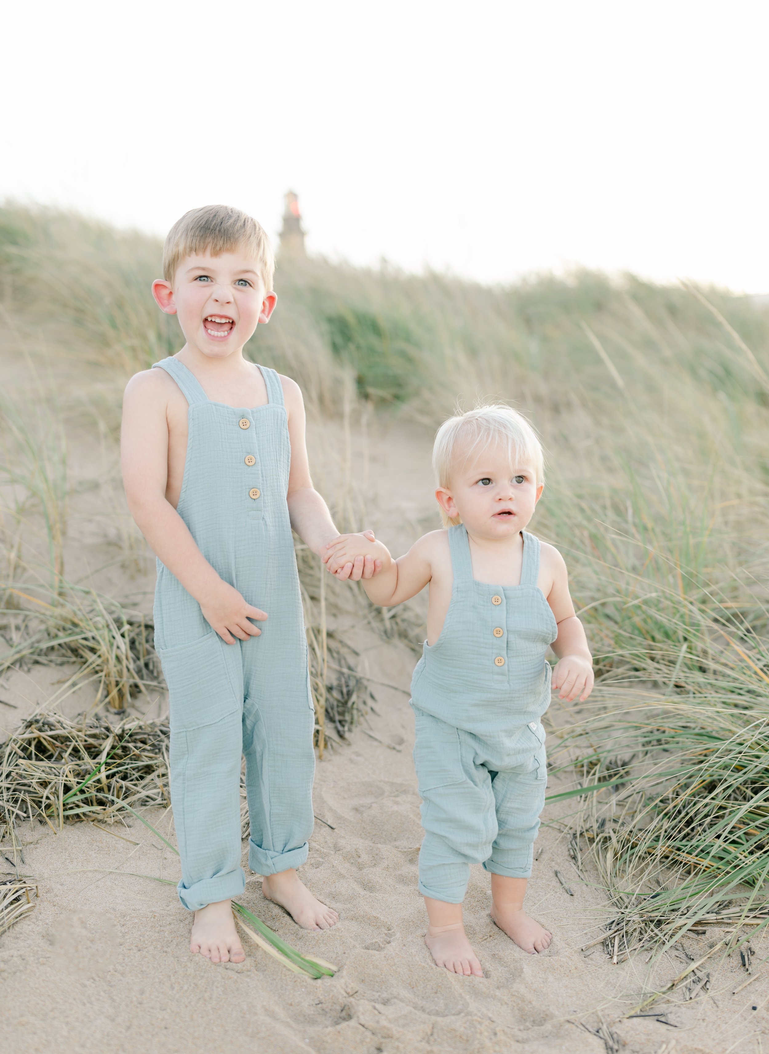 Two young children in matching light blue overalls standing on a sandy beach with grass.