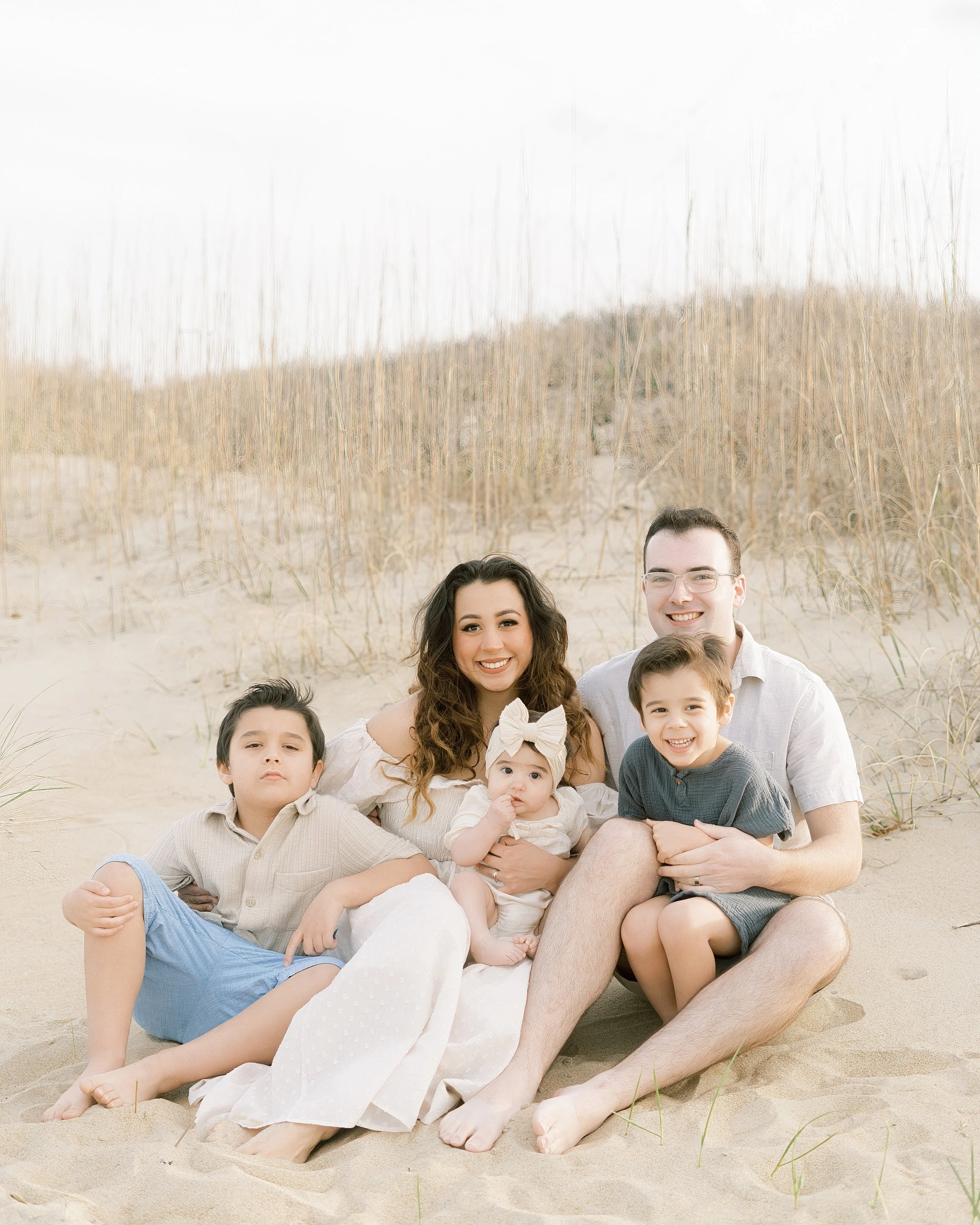 Family of five sitting on the sandy beach with tall grass and dunes in the background. The family includes a woman with long, curly hair, a man with glasses, a young boy in a gray shirt, a young boy in a beige shirt, and a young girl with a bow in he