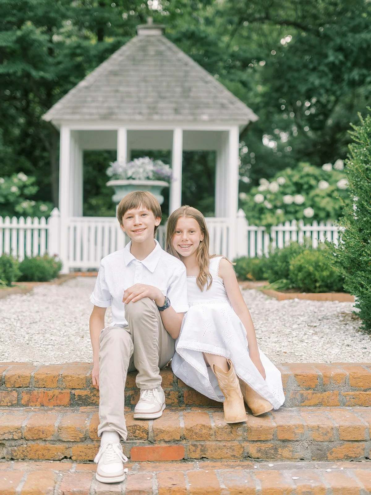 A boy and girl sitting on brick steps in front of a white gazebo with a shingled roof, surrounded by greenery.