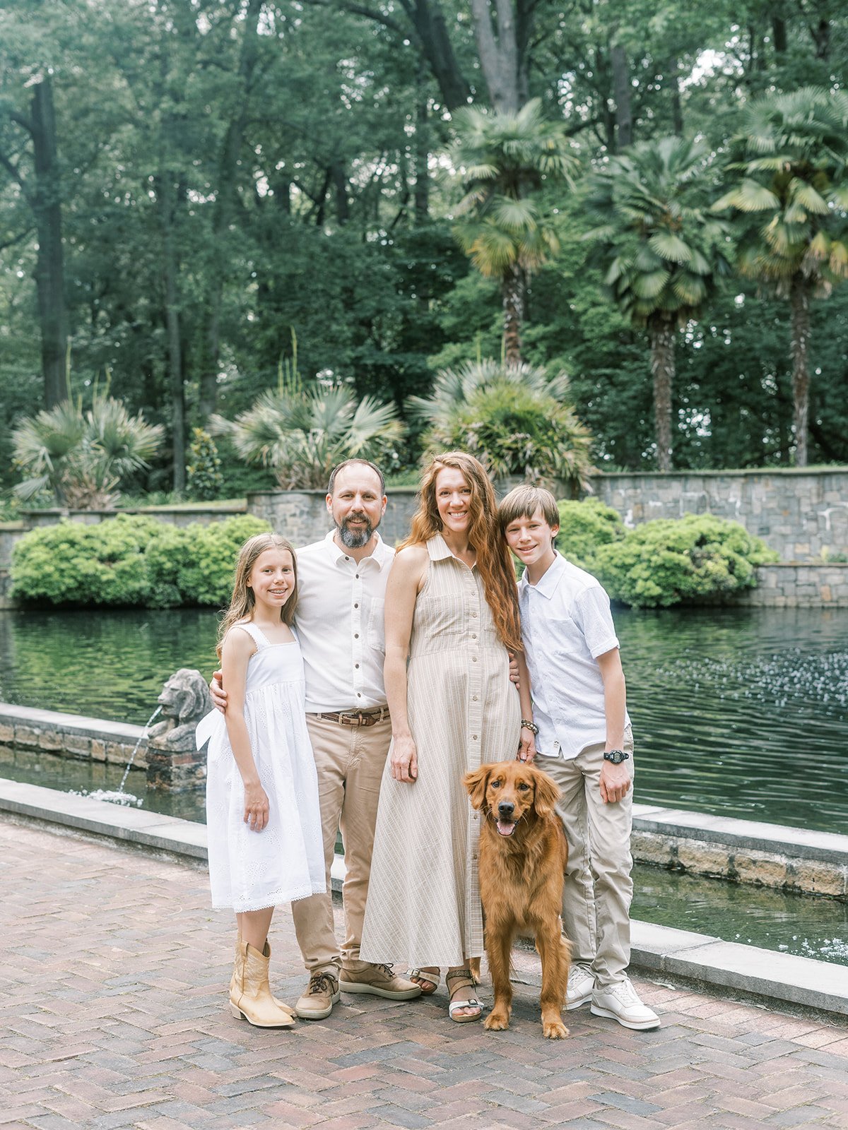 A family of four with a dog, standing outdoors by a pond in a lush, green park.