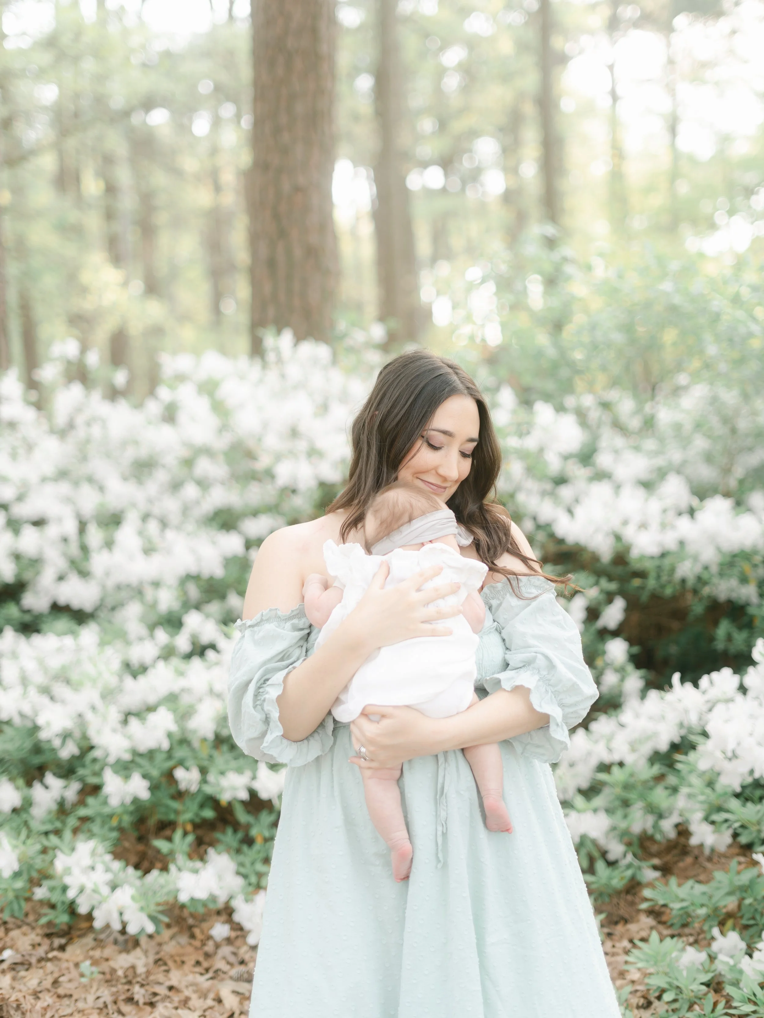 A woman holding a baby in a forest with white flowers and trees in the background.