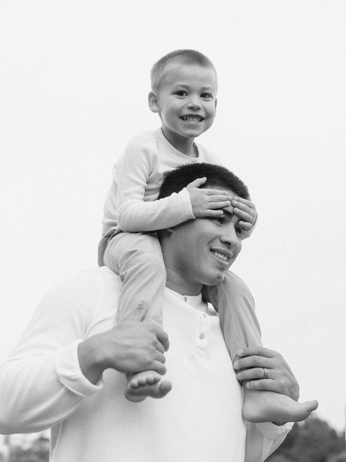 A man giving a piggyback ride to a smiling young boy on his shoulders, both looking toward the camera, outdoors against a plain sky.